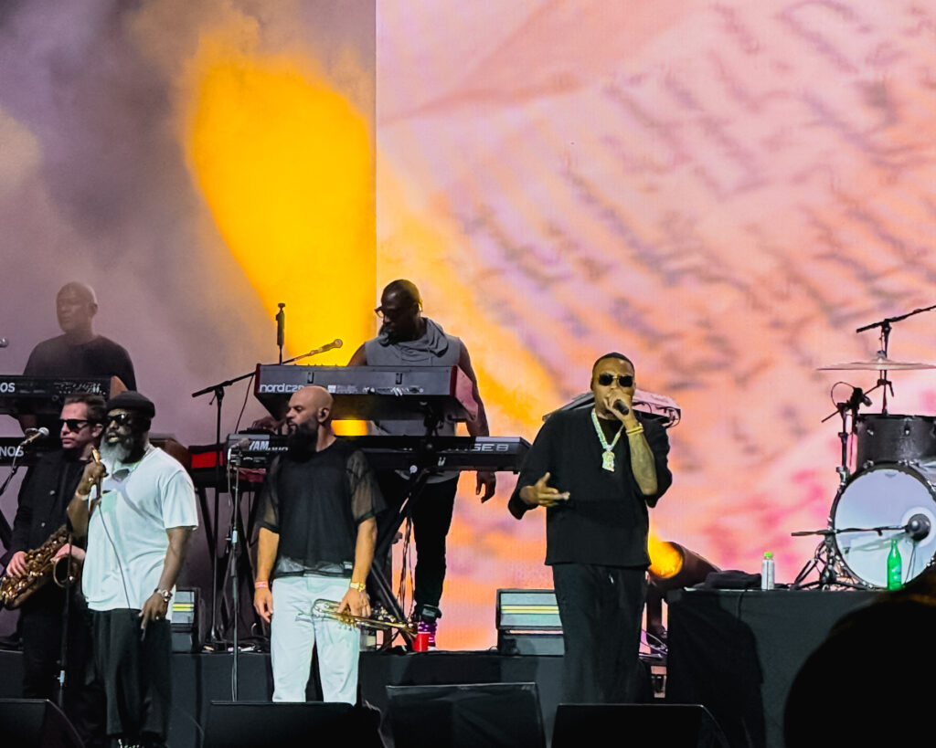 Nas-The-Roots-De-La-Soul-Great-Night-in-Hip Hop-05 – The 824 Journal Live concert stage with a singer at microphone in foreground and multiple musicians at keyboards and drums, with orange-pink backdrop smoke.