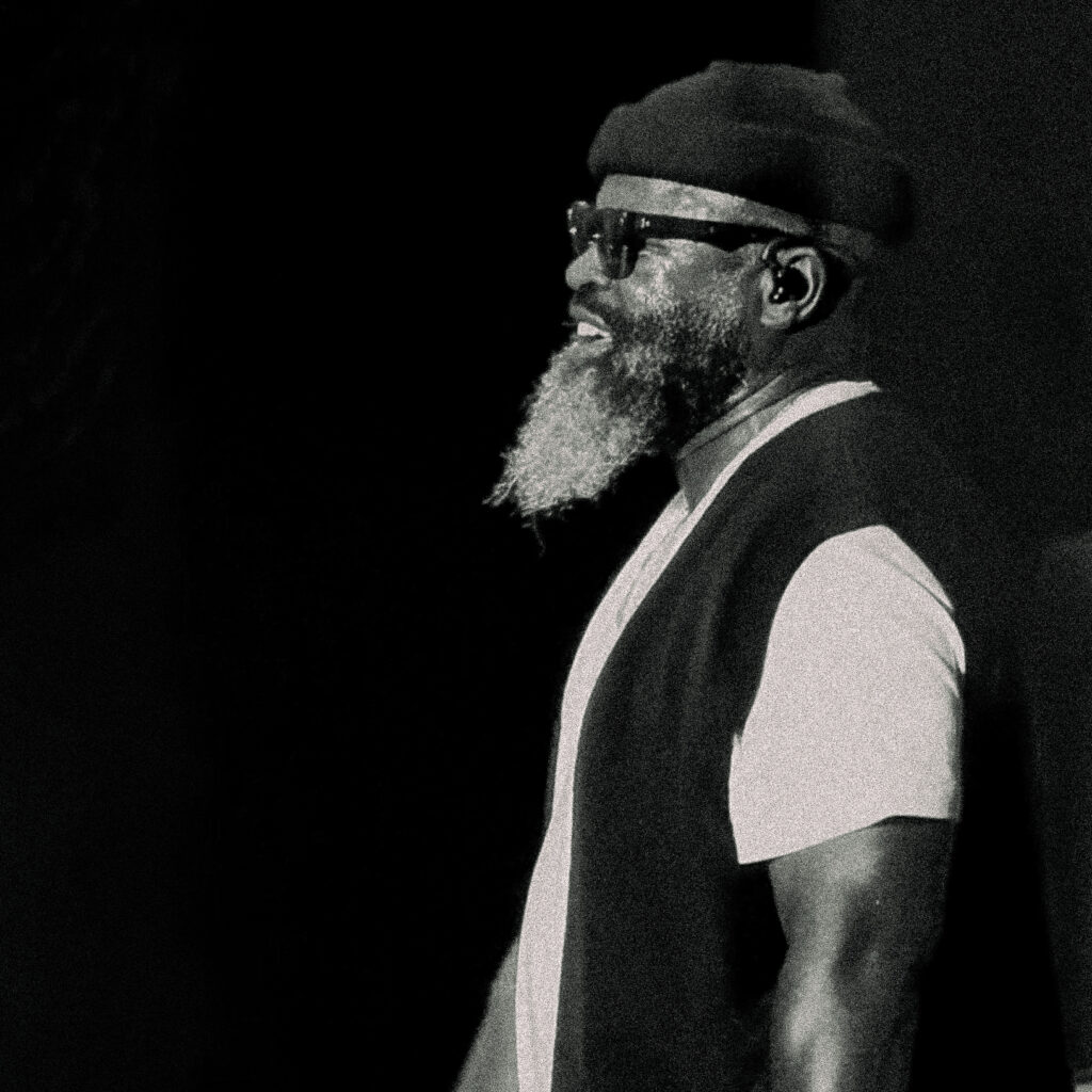 Nas-The-Roots-De-La-Soul-Great-Night-in-Hip Hop-06 – The 824 Journal Black-and-white portrait of a bearded man wearing a beret and glasses, smiling and facing left against a dark background.