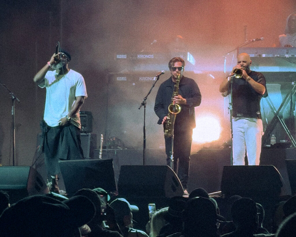 Screenshot – The 824 Journal Three musicians perform on a dimly lit stage: a singer left with a microphone, a saxophonist center, and a trumpeter right, with a cheering crowd in front.