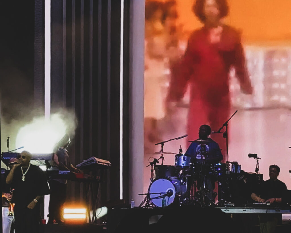 Screenshot – The 824 Journal Live concert stage with a male singer at left, drummer center, keyboardist at right; a large screen shows a blurry red-clad figure.