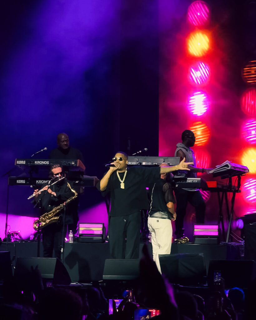 Screenshot – The 824 Journal Hip-hop artist in black with sunglasses sings into a microphone on stage, surrounded by keyboardists and a saxophonist amid purple-pink lights.