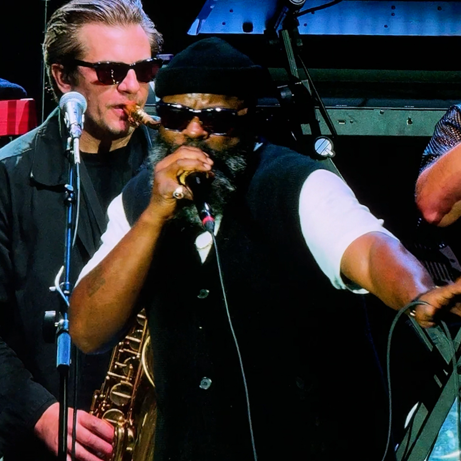 Zharmaine Boatman for The 824 Journal on iPhone 16 Pro – The 824 Journal Two male musicians on stage: a singer with a black vest and sunglasses, holding a microphone, and a saxophonist behind him wearing a hat and sunglasses.