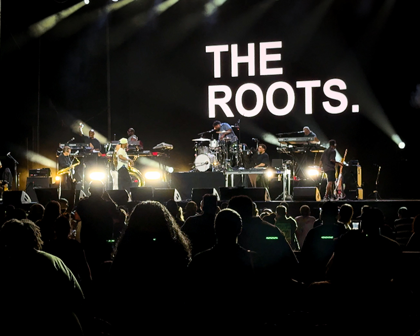 Screenshot – The 824 Journal The Roots performing on a dark stage with bright lights and a large THE ROOTS backdrop, audience silhouettes in the foreground.