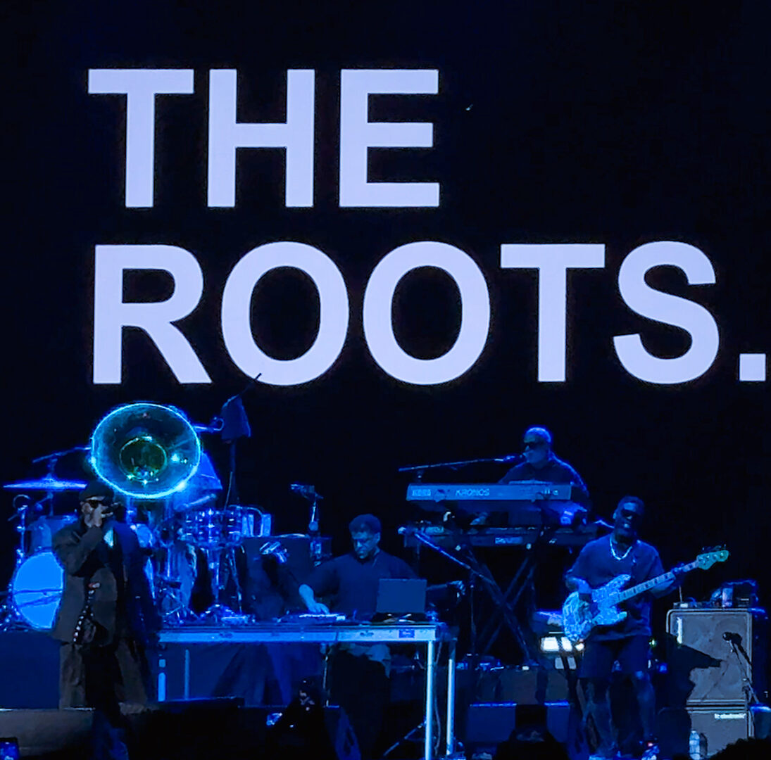 – The 824 Journal Live band on stage with a large 'THE ROOTS' backdrop, blue lighting and musicians playing keyboards, bass, and brass instruments.