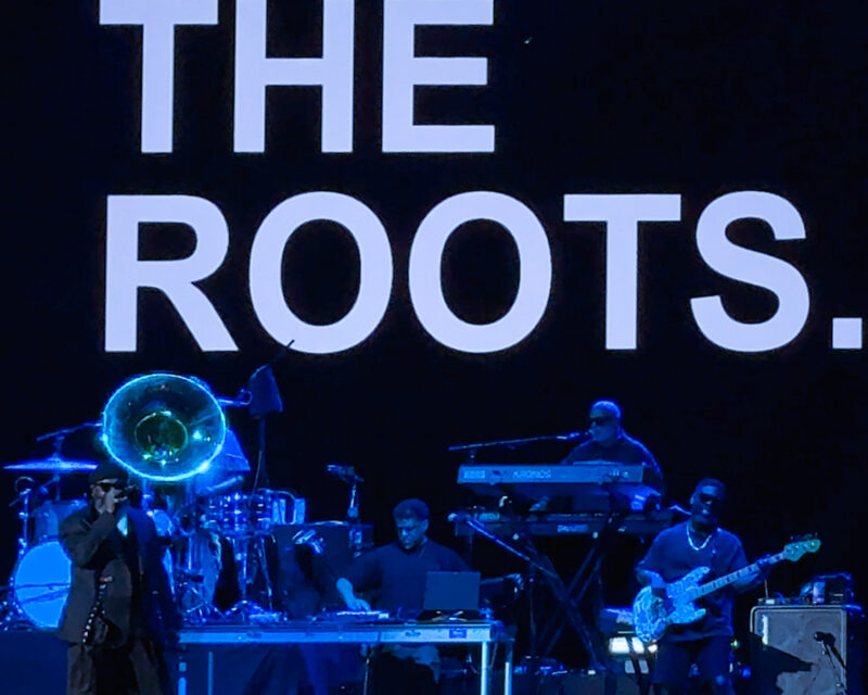Live band on stage with a large 'THE ROOTS' backdrop, blue lighting and musicians playing keyboards, bass, and brass instruments.