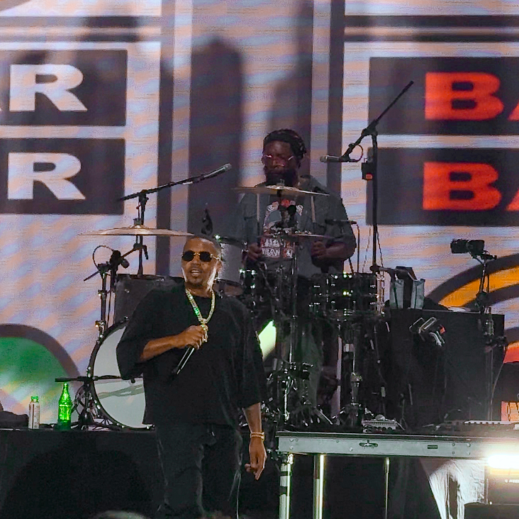 Screenshot – The 824 Journal Male performer in black with sunglasses sings into a microphone on stage, a drummer plays behind him with large LED screens in the background.