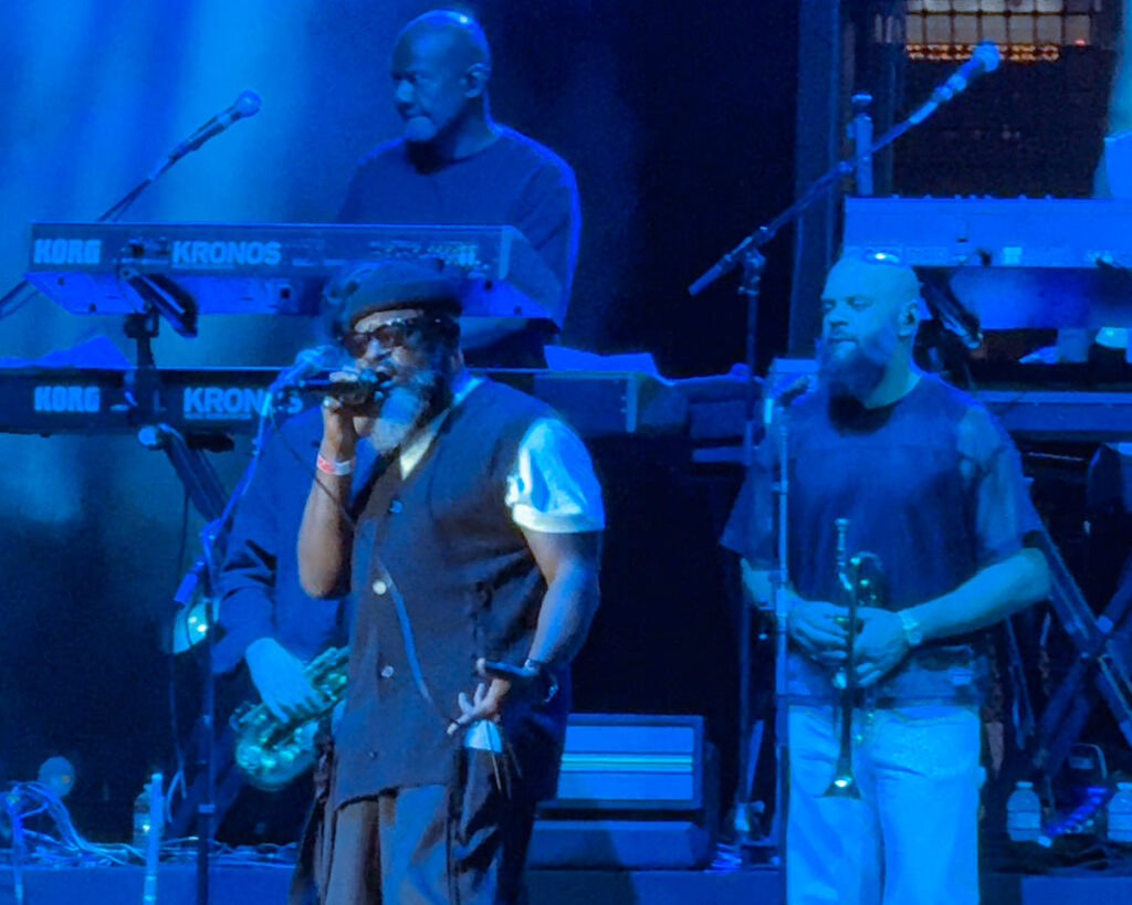 Screenshot – The 824 Journal Two male musicians perform on a blue-lit stage; one singer with glasses and hat in front, another man with a trumpet to his right, keyboardist in back.