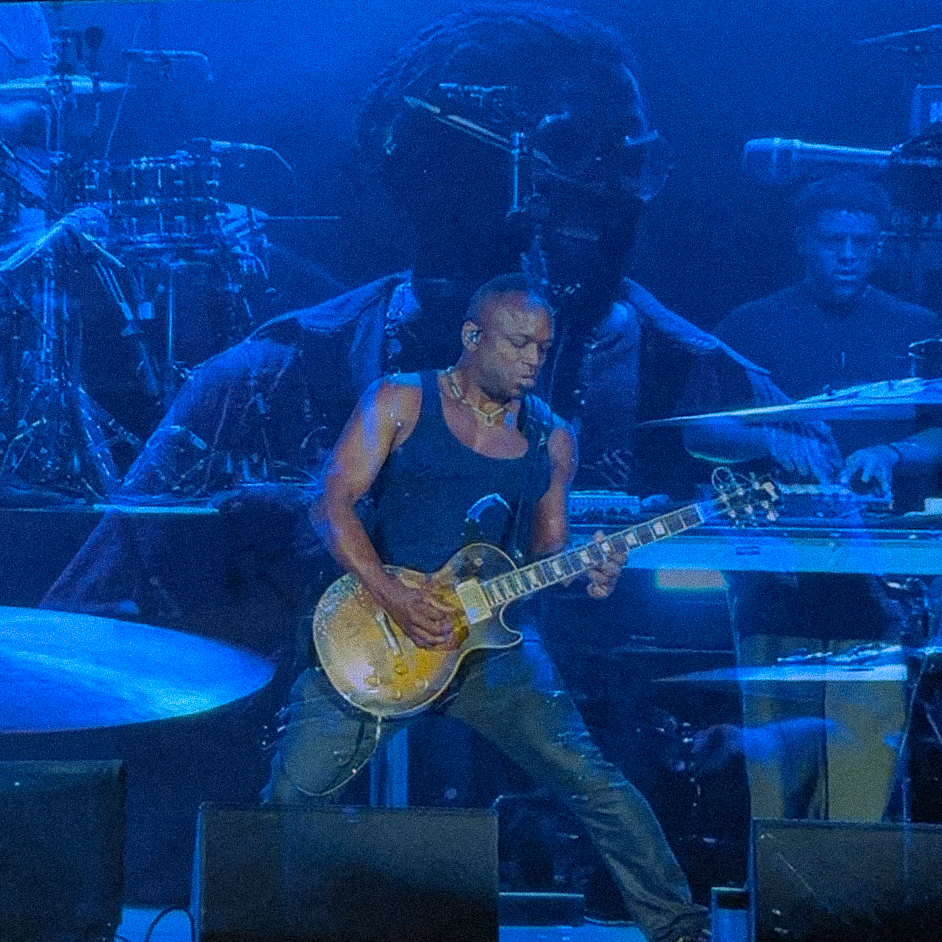Screenshot – The 824 Journal Guitarist performing on a blue-lit stage, wearing a blue sleeveless shirt and necklace, playing a gold electric guitar with band members in the background.