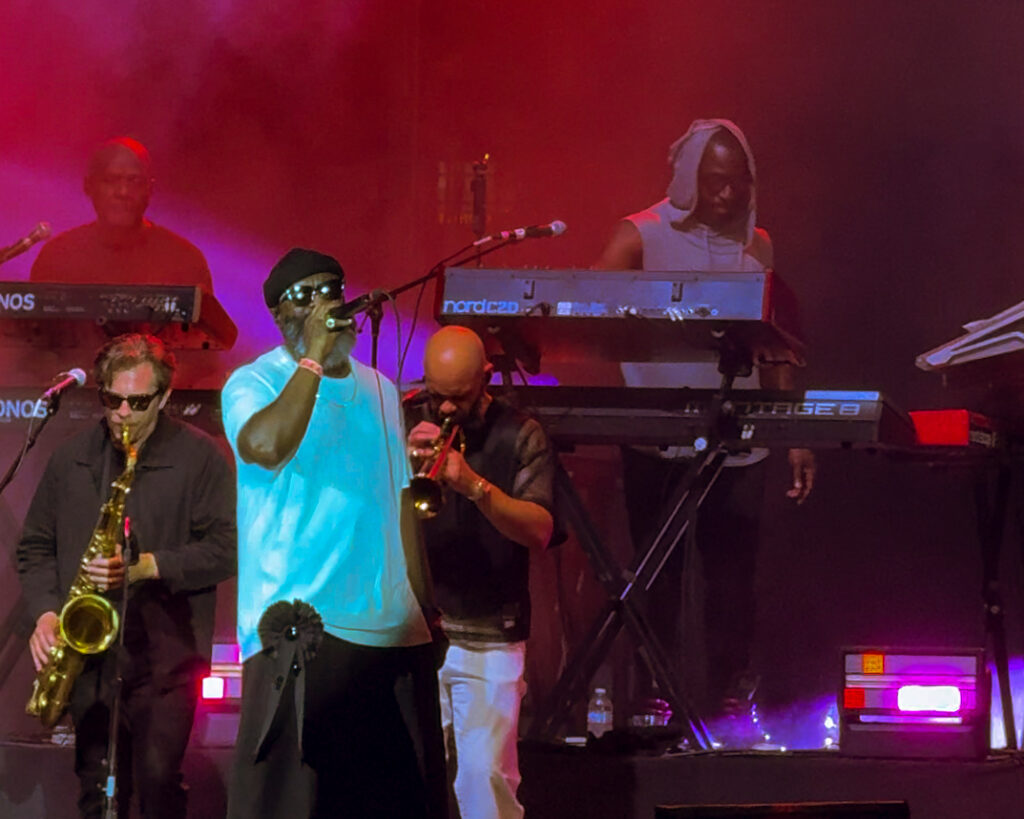 Screenshot – The 824 Journal A live band on stage performing under red and purple lights: a vocalist in a light blue shirt and black hat sings into a microphone, flanked by saxophonist and trumpeter; keyboard players behind them on stacked keyboards.