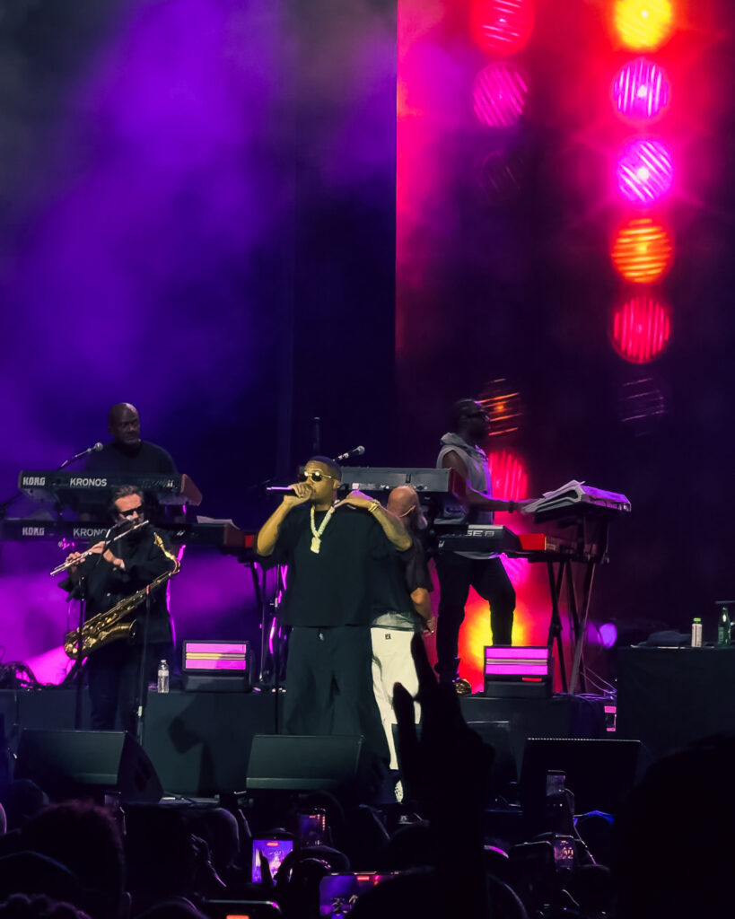 Screenshot – The 824 Journal Live concert scene with a rapper center stage wearing a black shirt and gold chain, performing among keyboardists and a saxophonist under purple and red lights.",