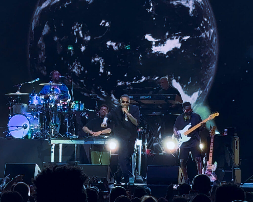 Screenshot – The 824 Journal Live concert on stage with a singer at center mic, guitarist to the right, drummer and keyboardist in back, Earth projection filling the screen behind.