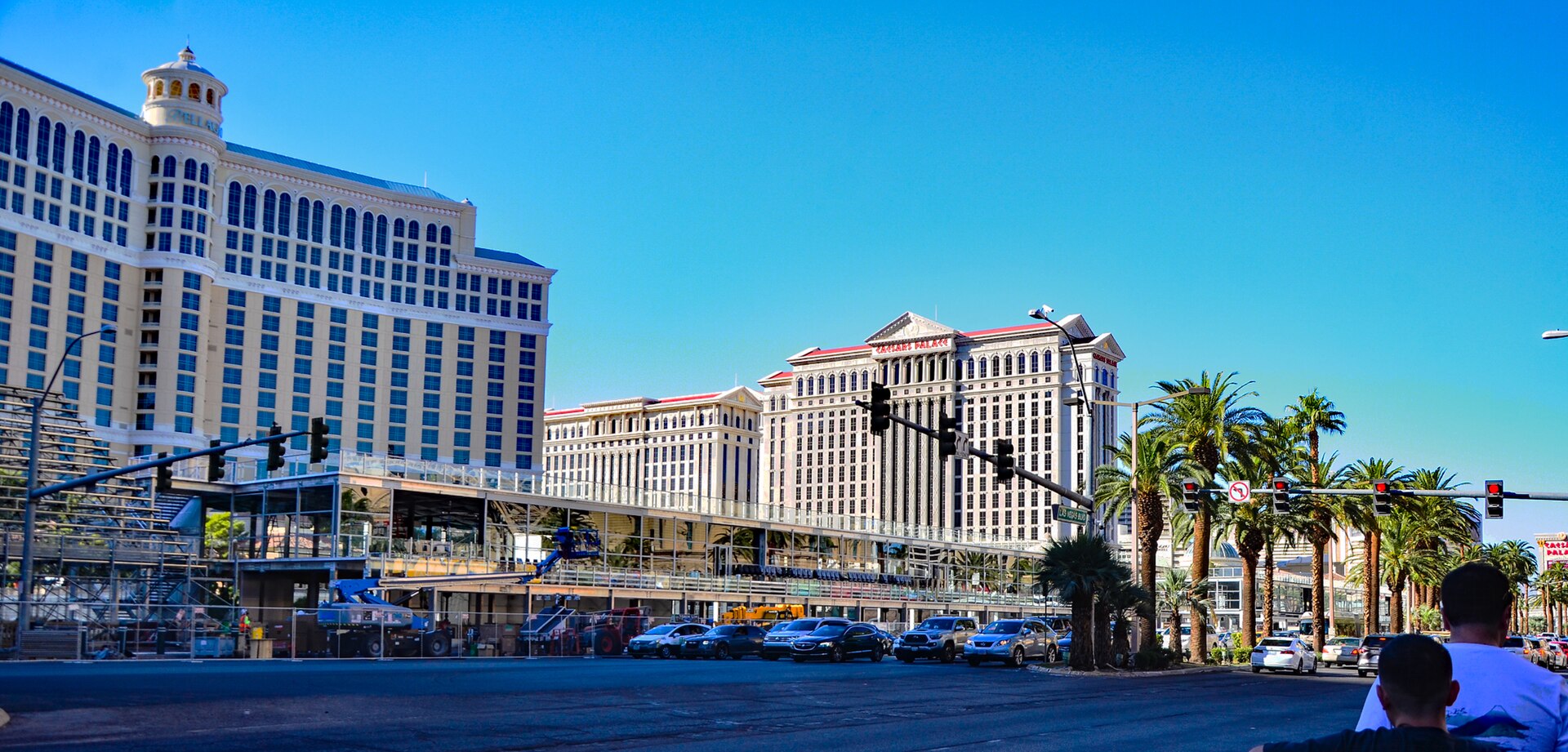 Las Vegas Strip scene with tall beige hotel towers, palm trees, and light construction fencing against a bright blue sky.