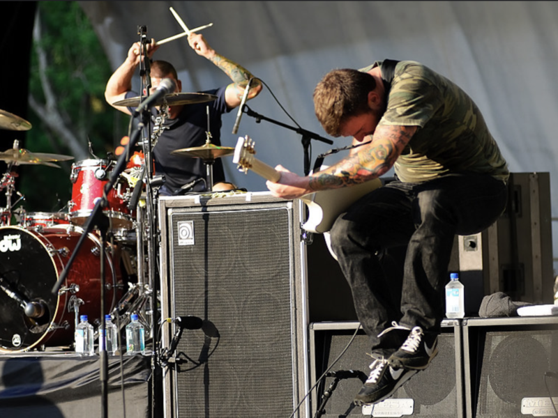 New Found Glory guitarist Chad Gilbert performing on stage beside the drum kit.