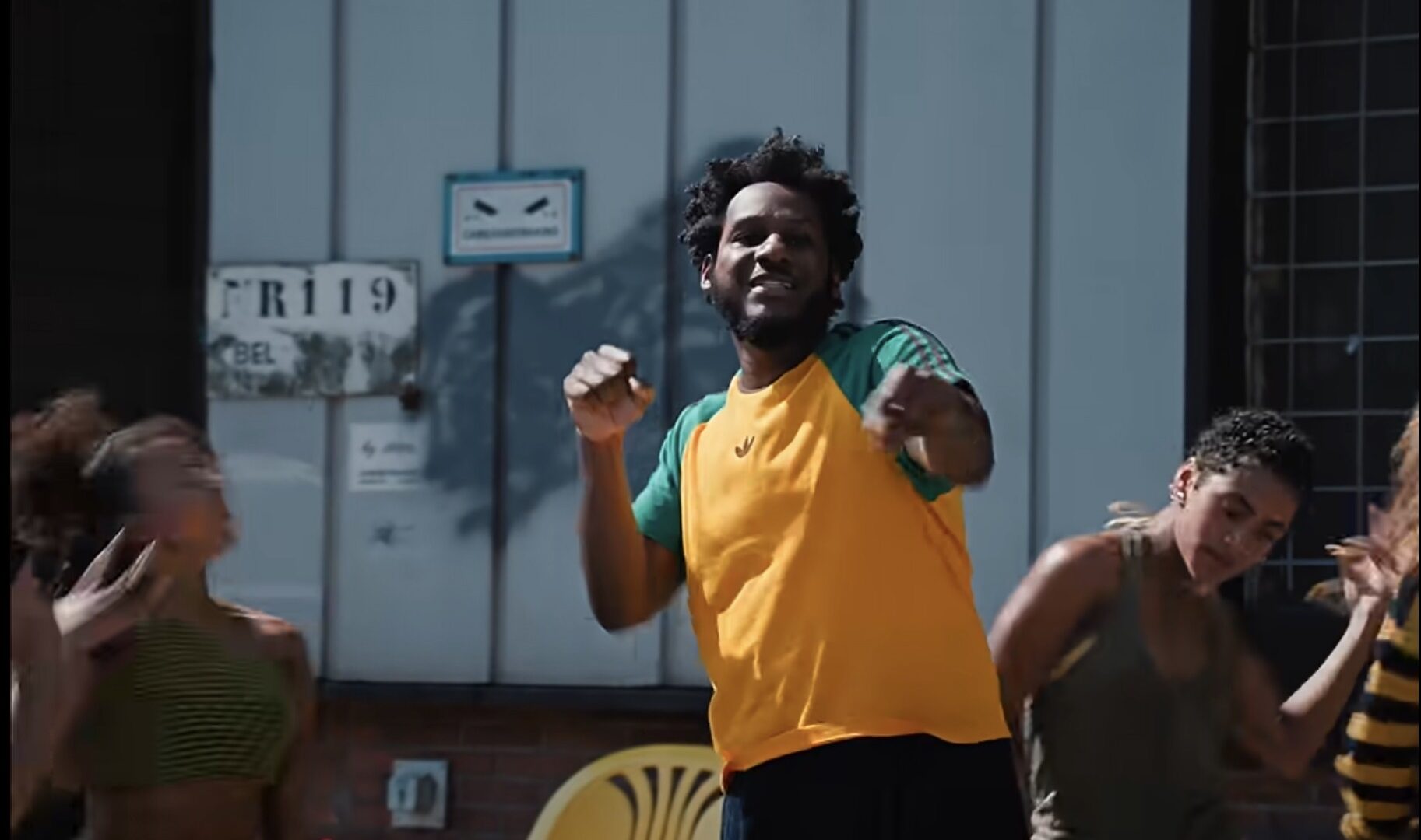Leon Bridges Video Still – The 824 Journal A man in a yellow and green shirt dances in the foreground with others nearby outdoors.