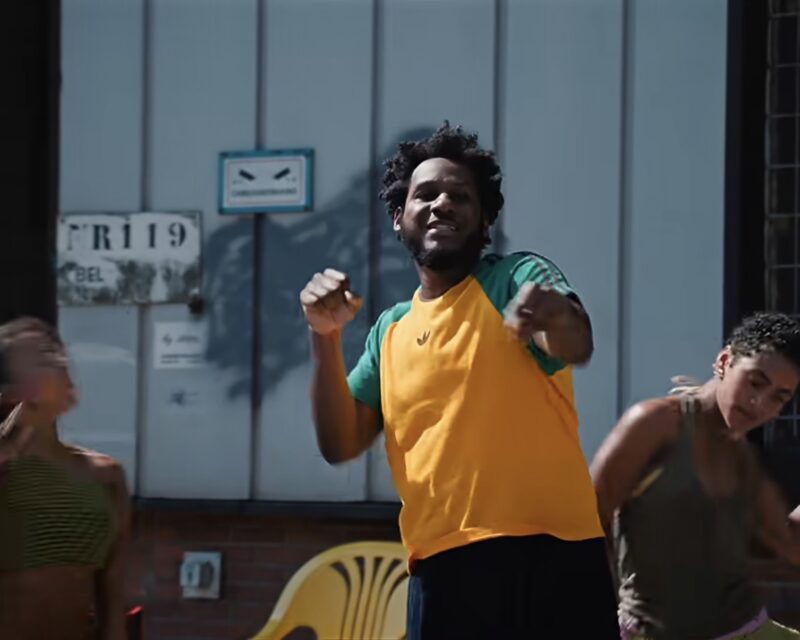 A man in a yellow and green shirt dances in the foreground with others nearby outdoors.