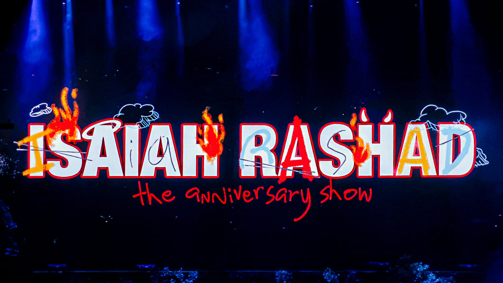 Neon sign with large white-outlined letters spelling 'ISAAH BASHIAD' over a dark stage, orange flames painted across the letters, and red handwritten text below reading 'the Anniversary Show'