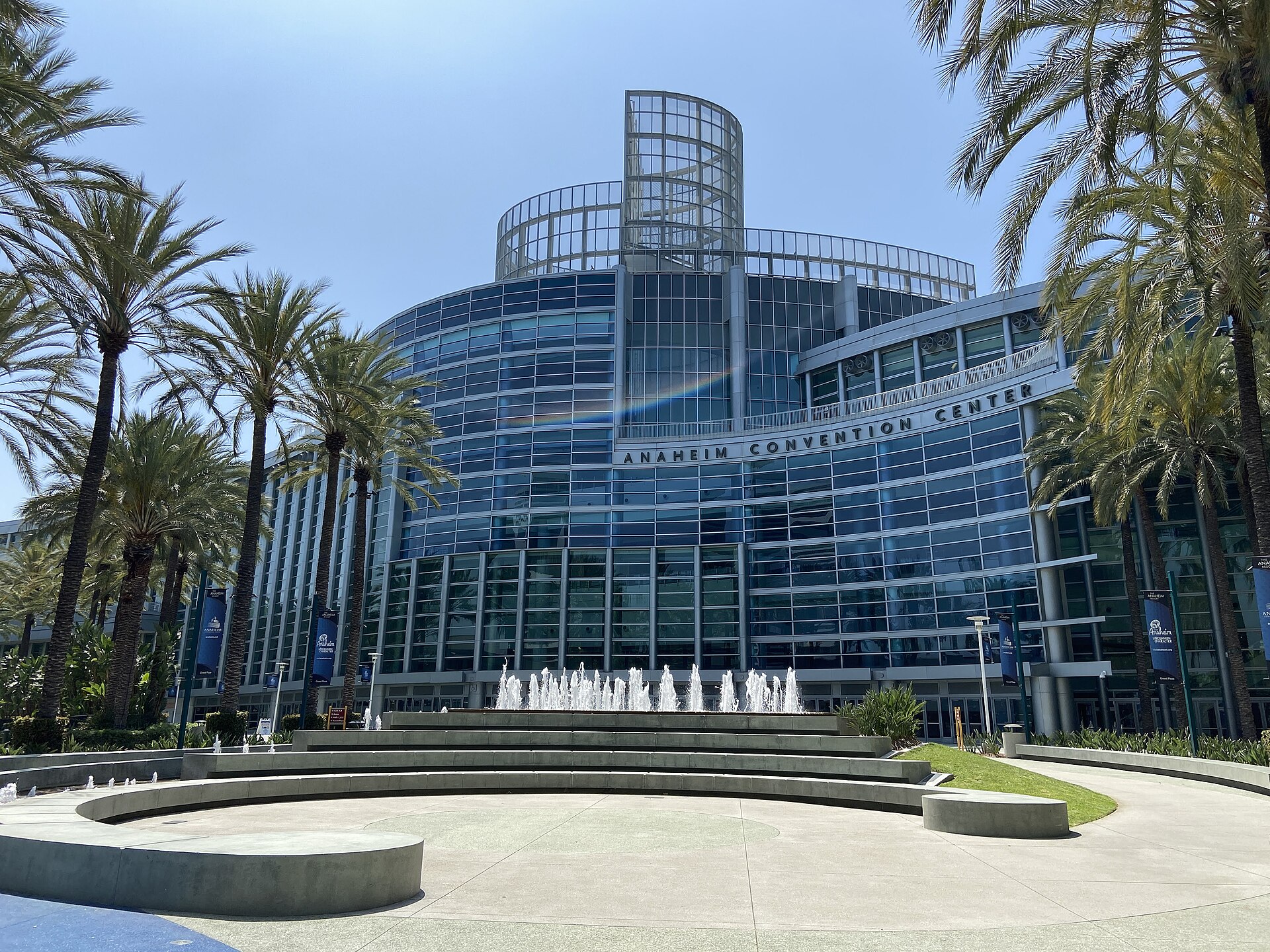 The curved blue glass front of the Anaheim Convention Center behind a plaza fountain, framed by palm trees under a clear sky