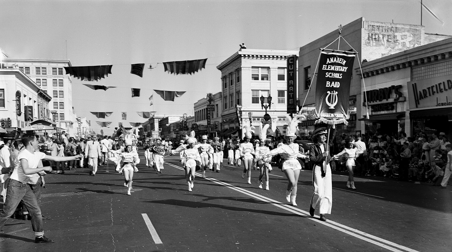 Black-and-white photo of a marching band parading down a downtown Anaheim street under Halloween bunting, with crowds lining the sidewalks