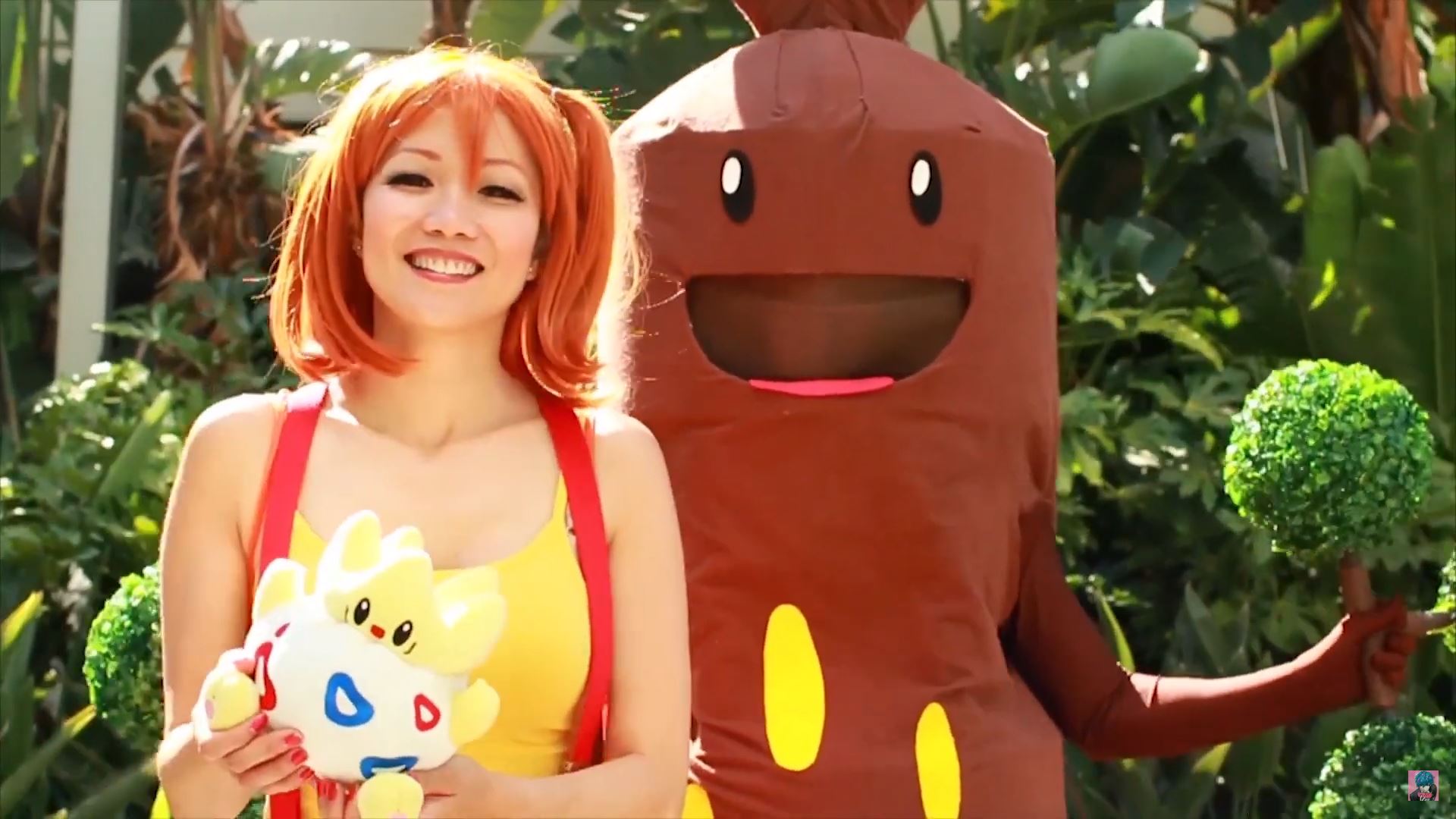 Smiling woman in a yellow outfit holding a colorful plush toy, with a person in a brown mascot suit beside her outdoors.