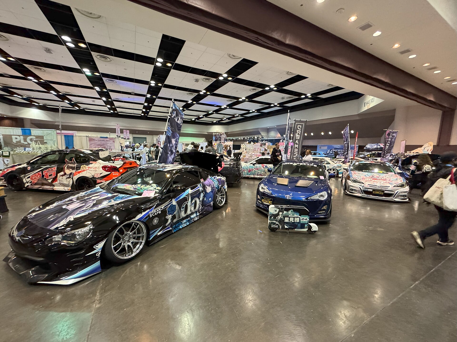 Rows of itasha cars wrapped in colorful anime character artwork parked inside a convention hall
