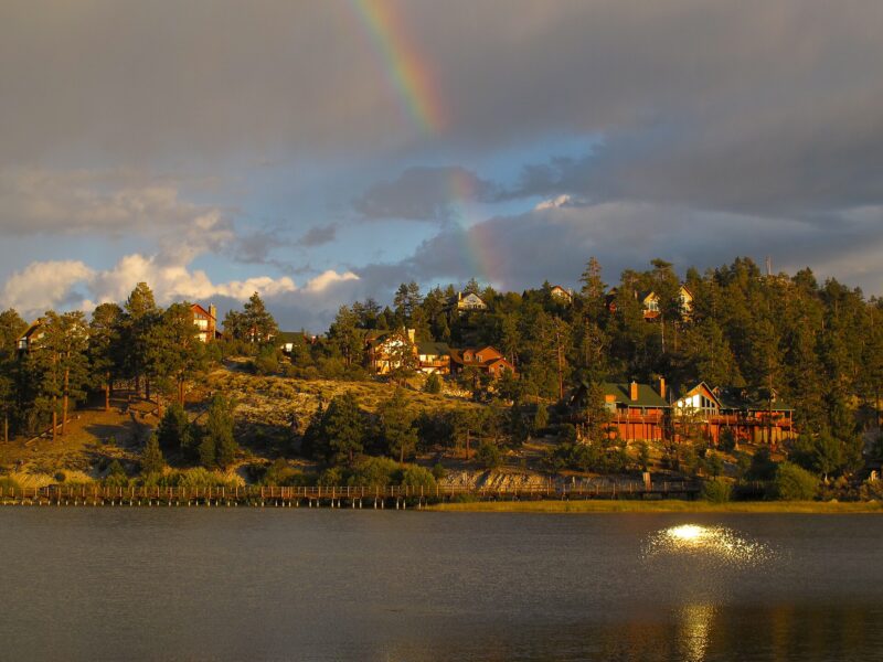 A rainbow over pine trees and lakeside homes on the shore of Big Bear Lake under dark clouds