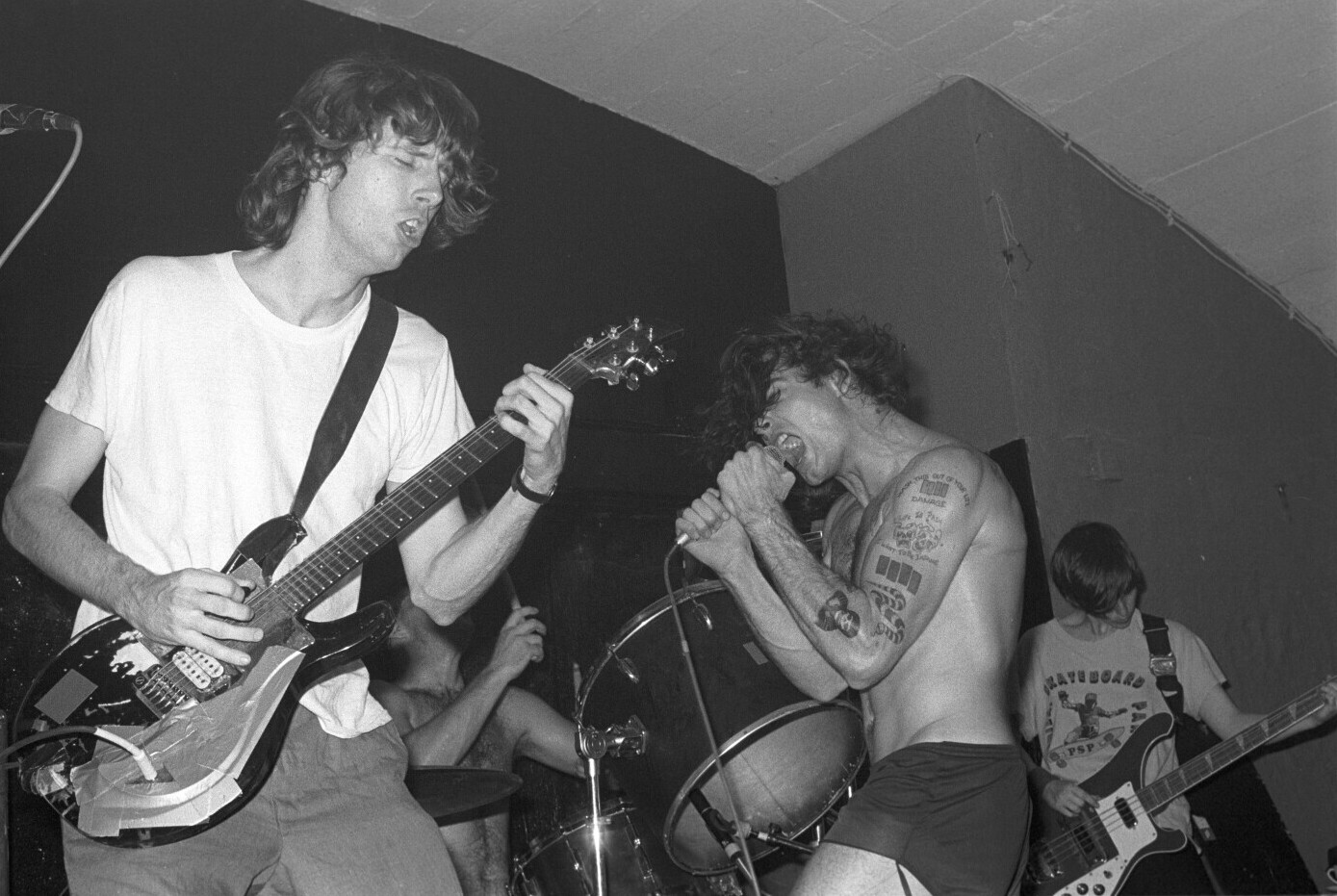 Black-and-white photo of Black Flag playing a small club stage, a guitarist in the foreground and the singer shirtless at the microphone