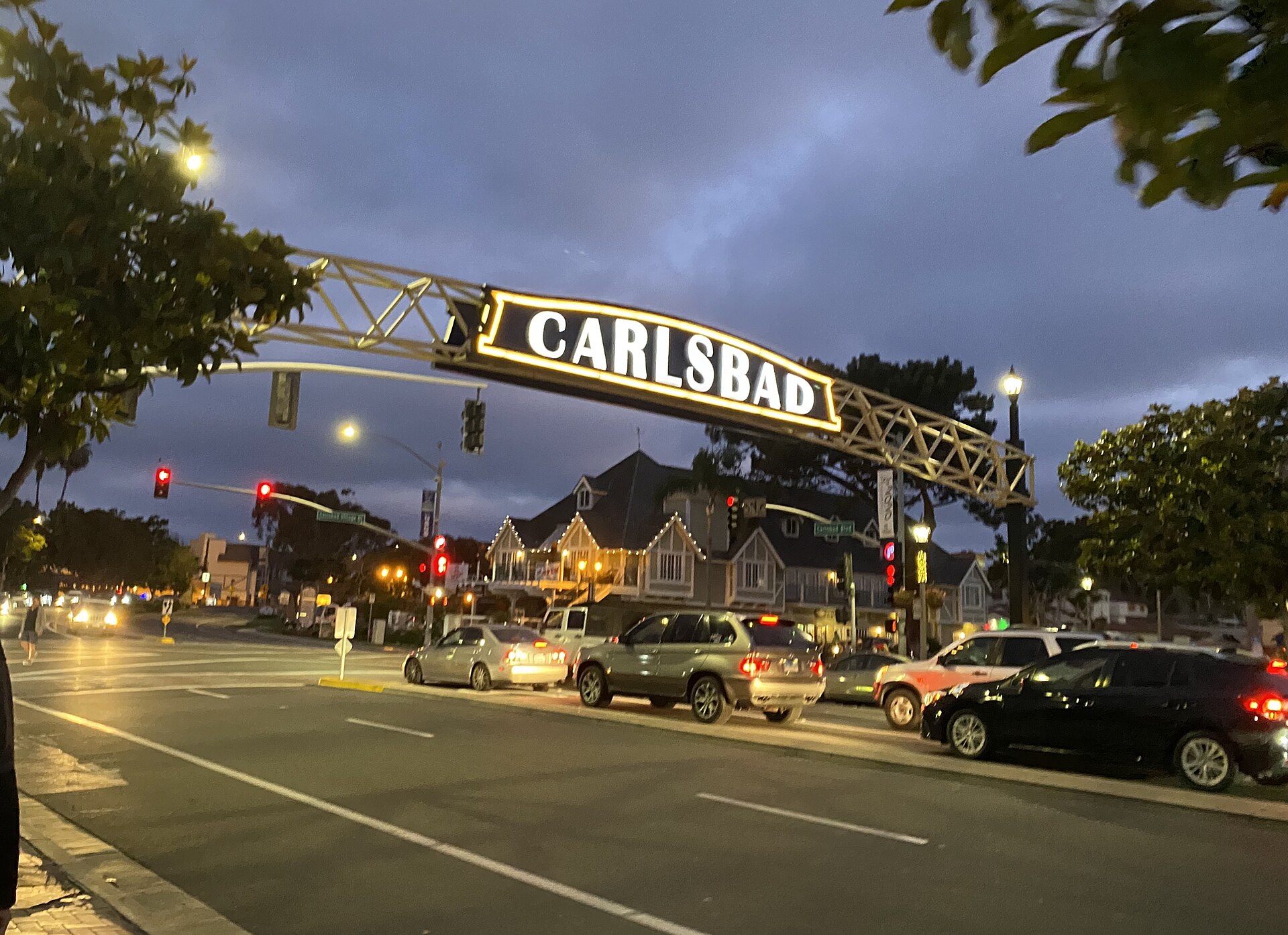 The arched Carlsbad sign lit up over Carlsbad Boulevard in the village at dusk, with parked cars and trees along the street