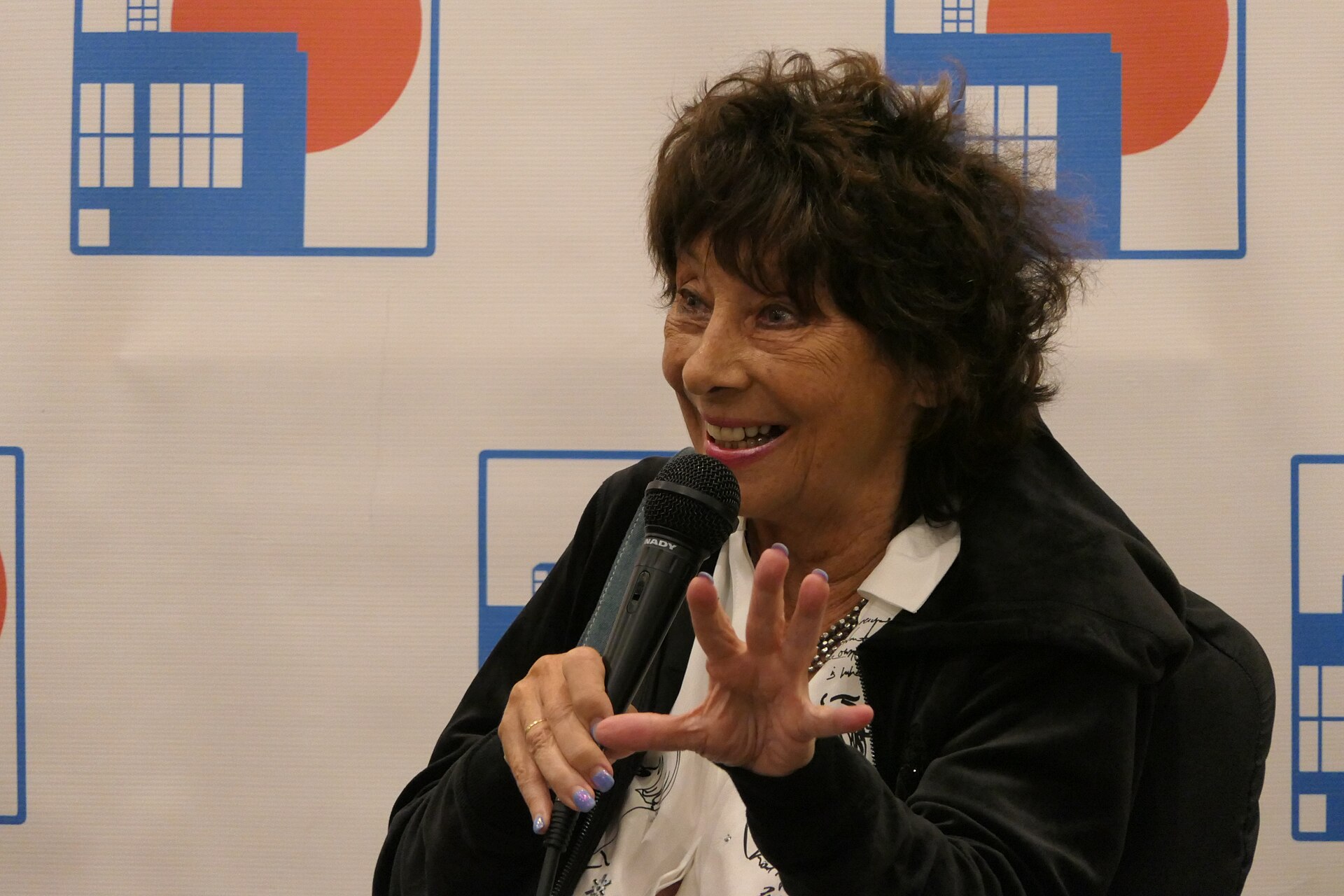 Carole Ann Ford smiling and gesturing as she speaks into a microphone in front of a backdrop printed with blue police box graphics