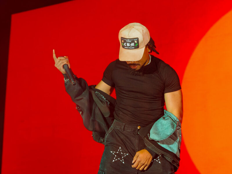 Chance the Rapper performing at the Hollywood Palladium in a beige cap with a denim jacket tied at his waist, microphone raised against a red-orange backdrop.