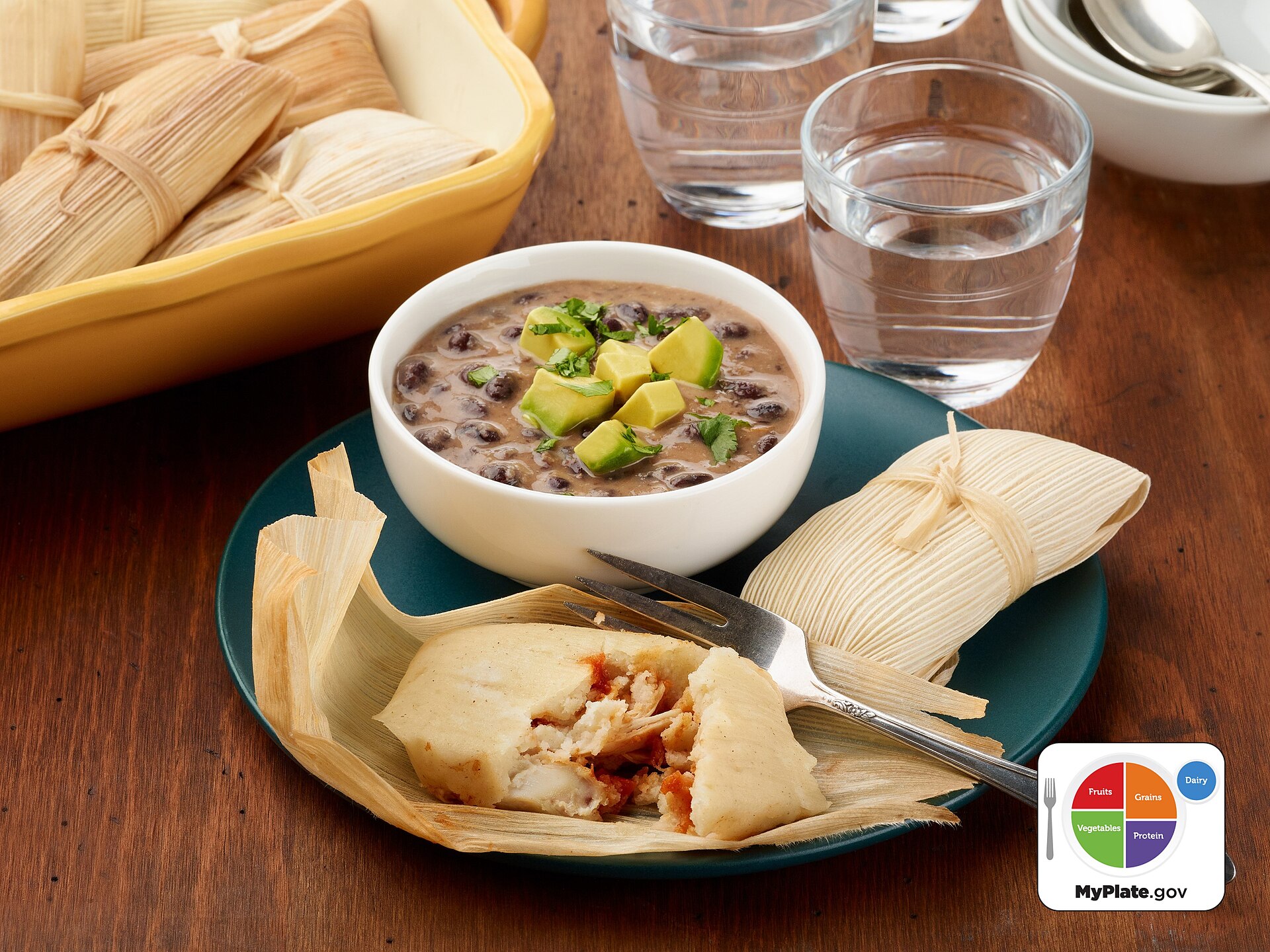 Chicken tamales on a plate beside their corn husk wrappers, with a small bowl of salsa and glasses of water on a wooden table