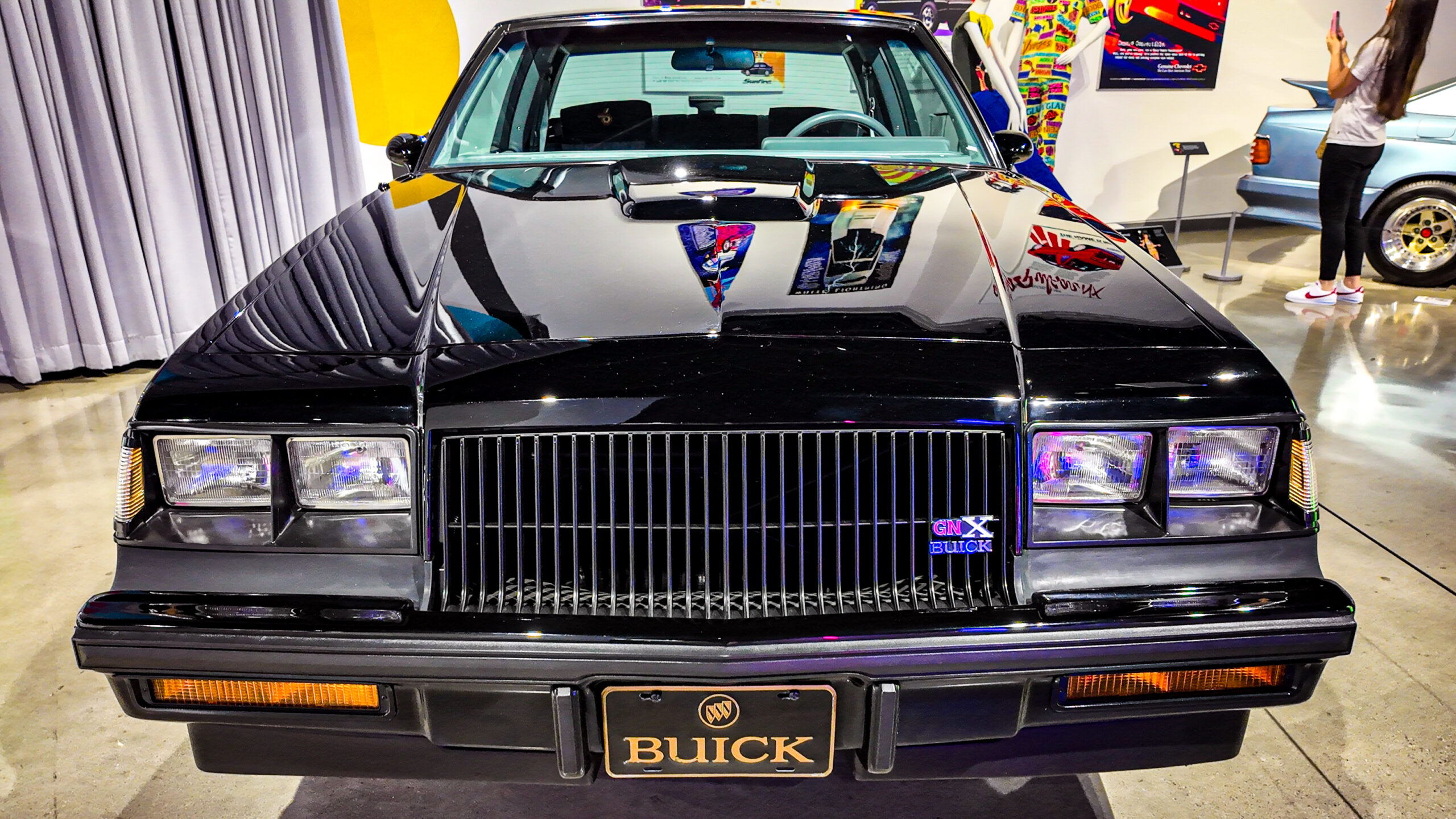 Front view of a glossy black Buick GNX on display in a showroom with a bold grille and rectangular headlights near the camera edge to the left and right margins, reflecting surrounding posters and lights. (Informative)