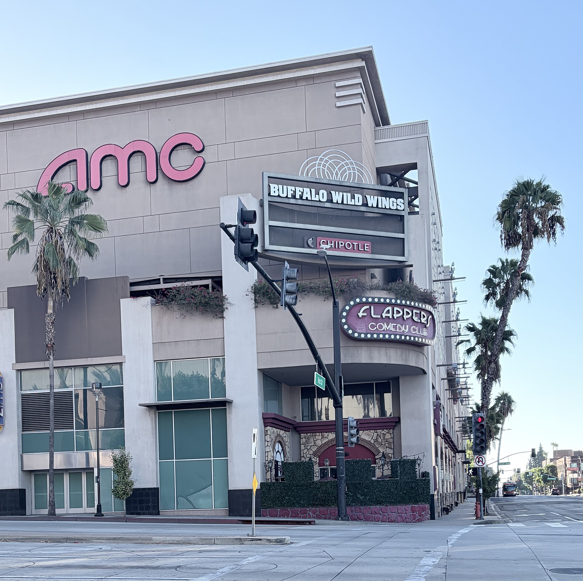 A street corner in downtown Burbank with the AMC Burbank 16 theater sign, the Flappers Comedy Club marquee, and tall palm trees under a blue sky