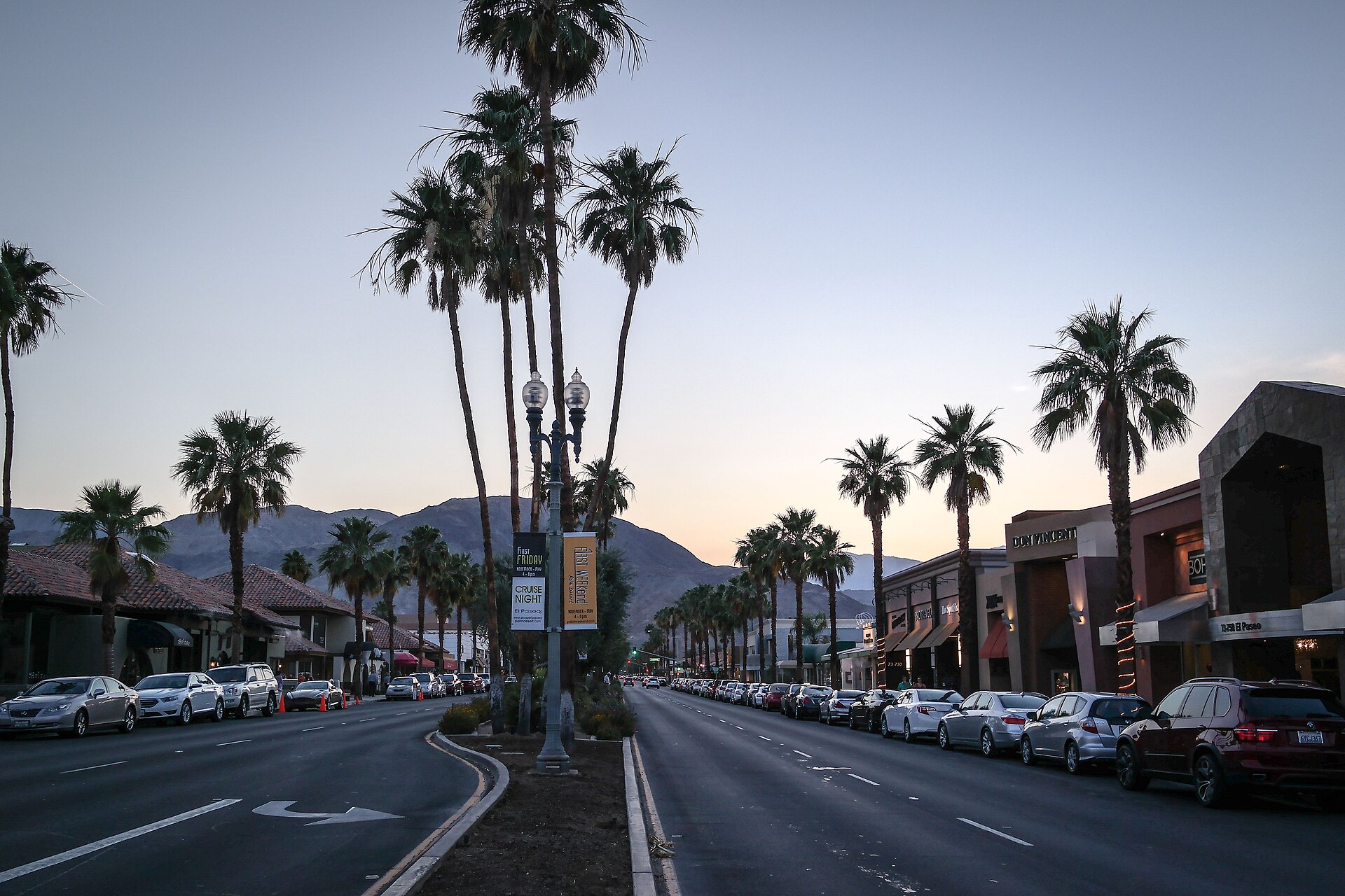 El Paseo in Palm Desert at dusk, lined with tall palm trees, shops, and parked cars, with mountains in the distance