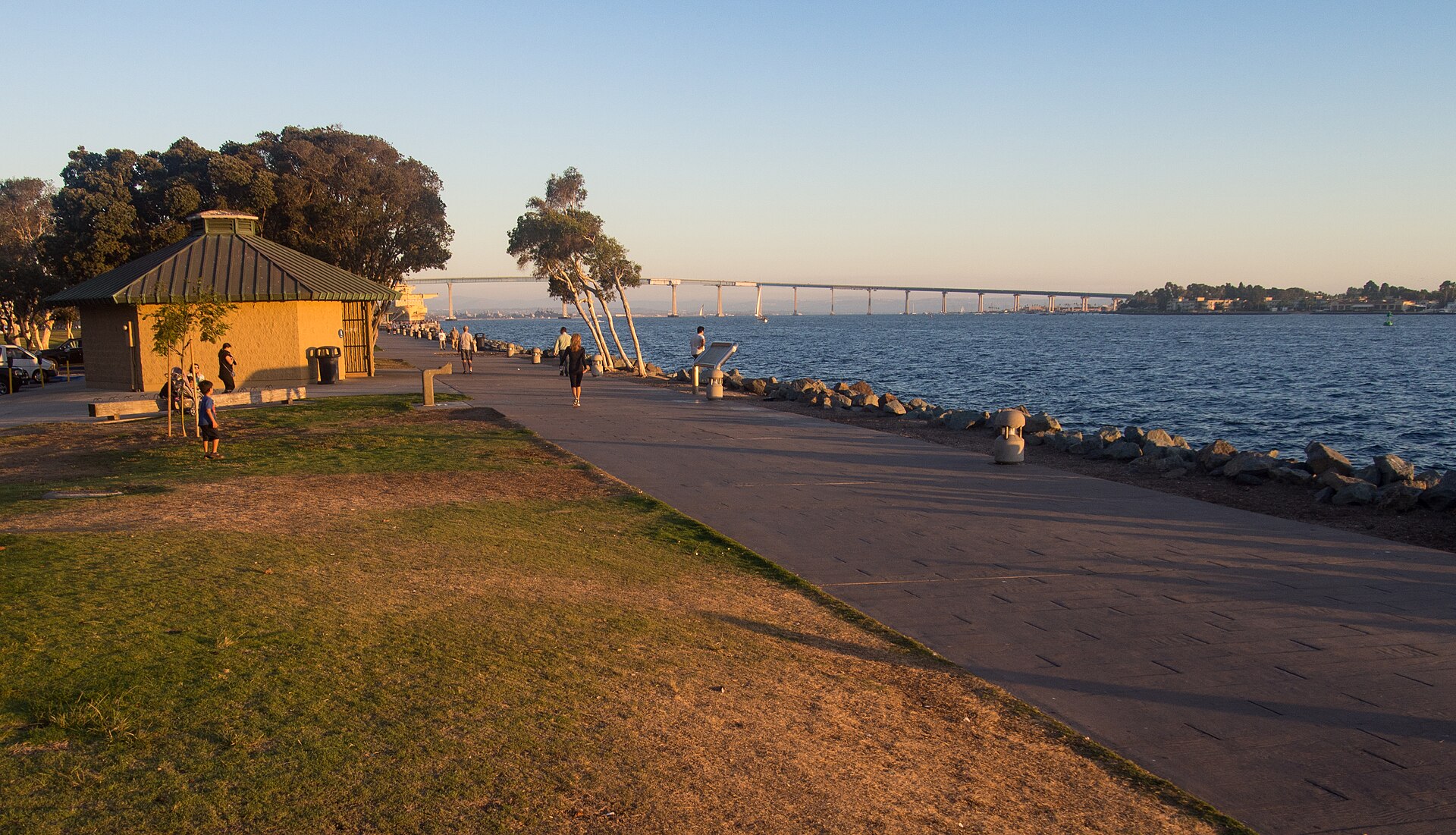 A waterfront path and lawn at Embarcadero Marina Park North at sunset, with San Diego Bay and the Coronado Bridge in the distance