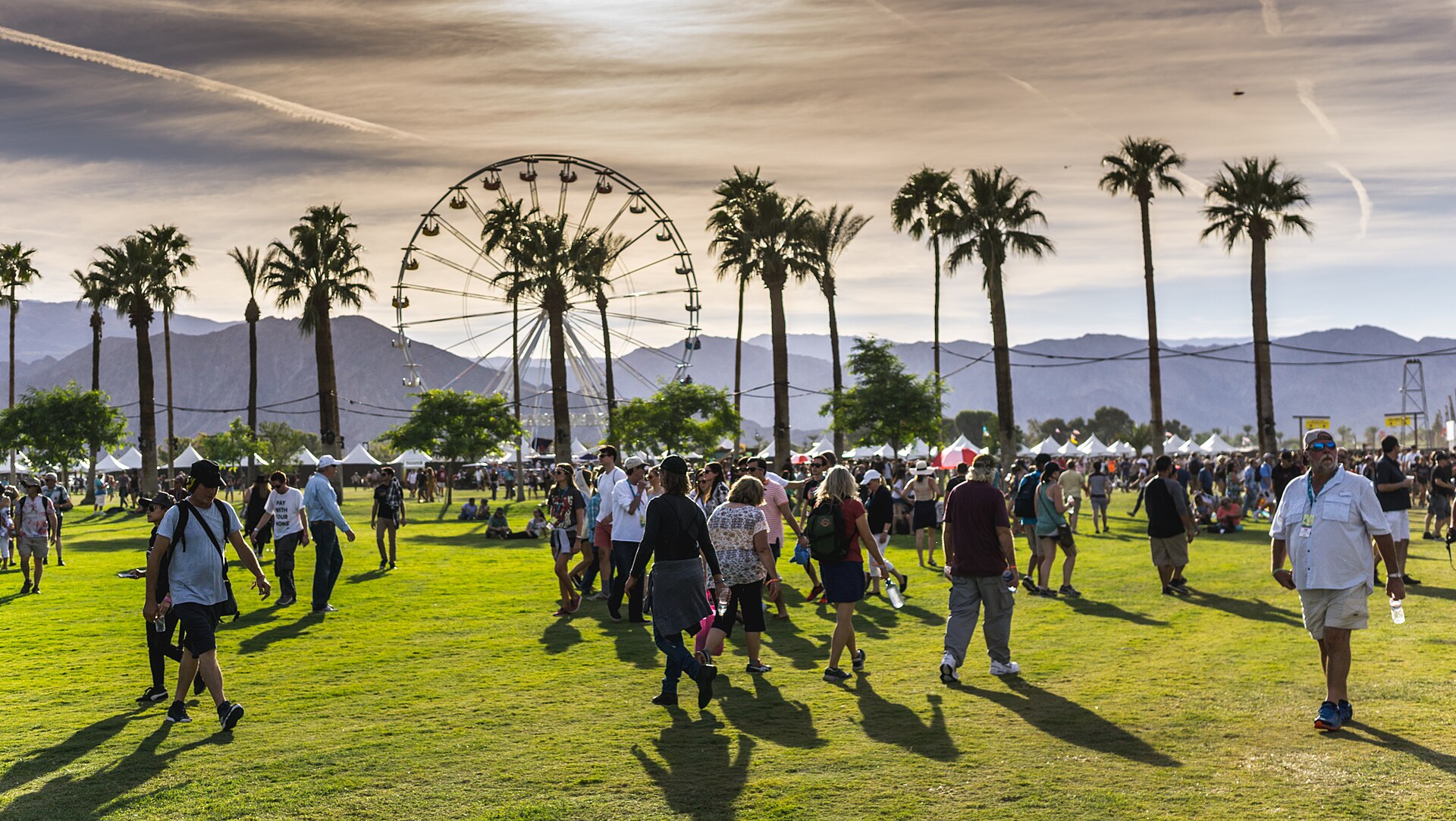 Festivalgoers walking across the green lawn at the Empire Polo Club, with rows of palm trees, a Ferris wheel, and desert mountains behind