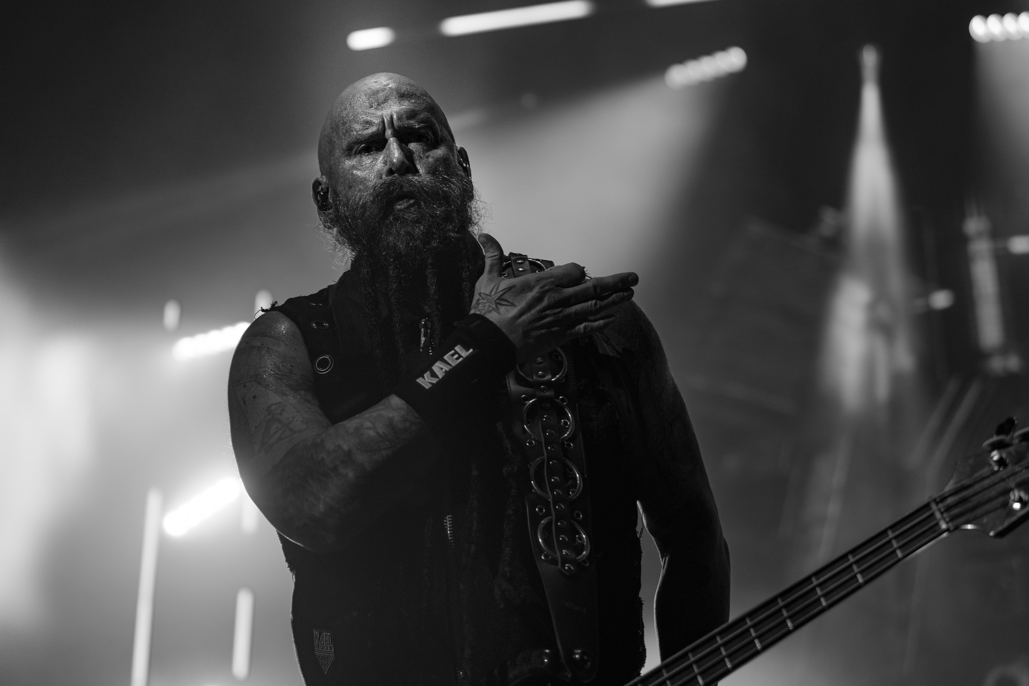 Five Finger Death Punch bassist Chris Kael performing at F&M Bank Amphitheater in Long Beach, hand pressed to his chest under bright stage lights, in black and white.