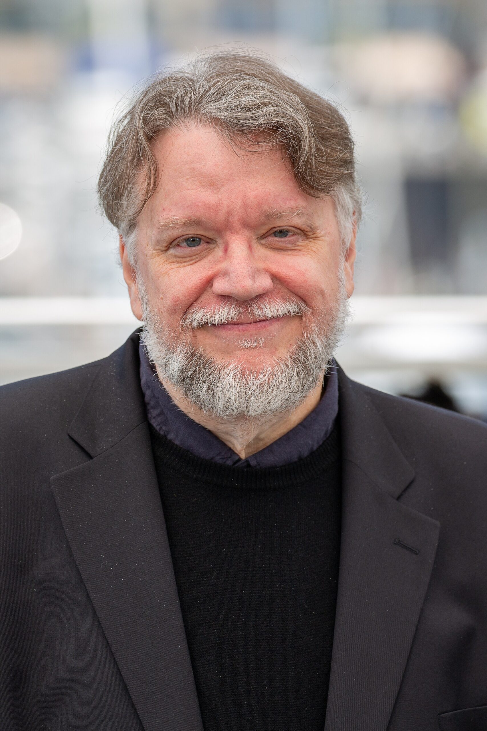 Filmmaker Guillermo del Toro smiling in glasses, a black sweater, and a dark blazer at a photocall