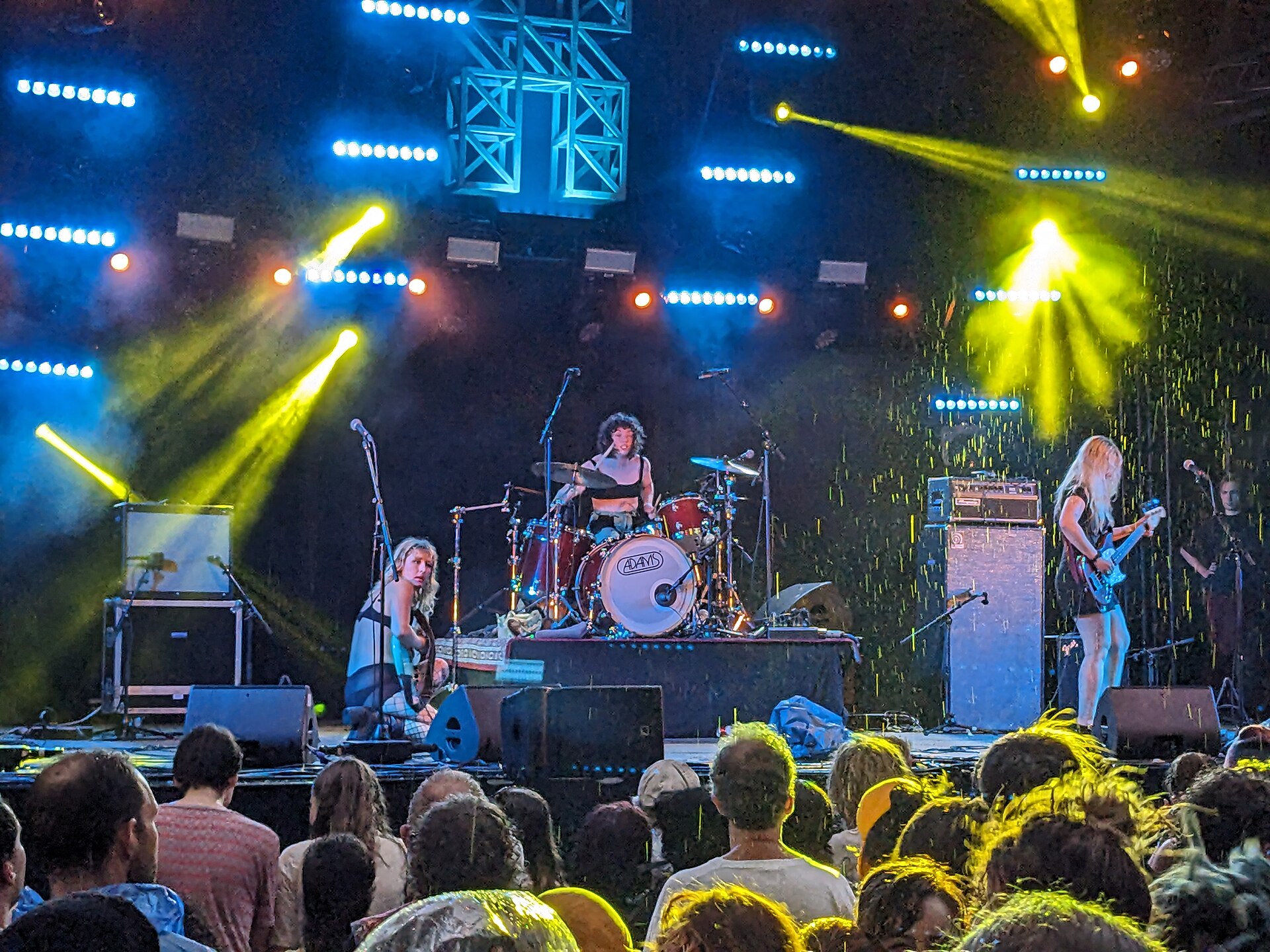 Lambrini Girls performing on an outdoor festival stage under yellow and blue lights, seen over a crowd