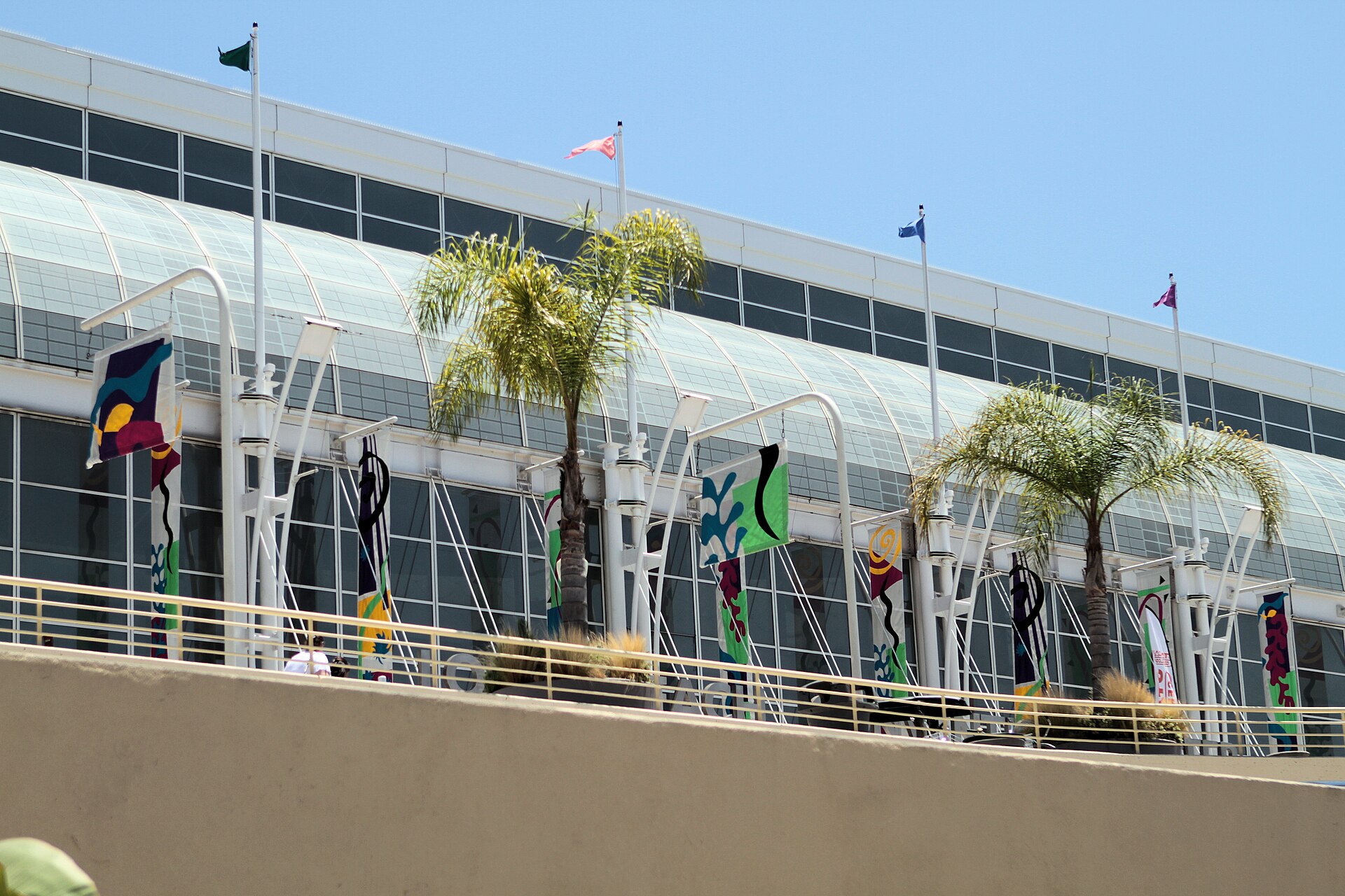 The glass facade of the Long Beach Convention Center with flags and palm trees along the upper terrace under a blue sky