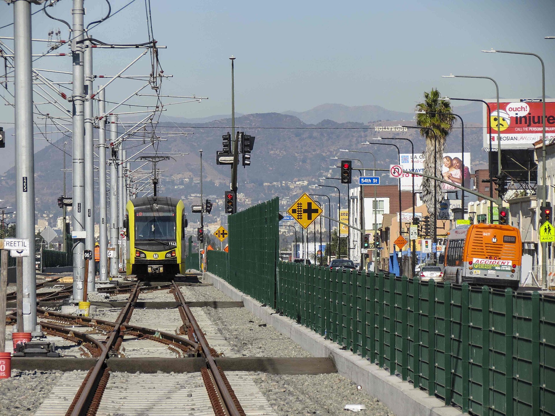 A Metro K Line light rail train running in the median of Crenshaw Boulevard, with shops and palm trees along the street and hills in the distance