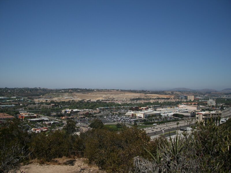 A wide view across central Mission Valley in San Diego from a brushy hillside, with buildings and freeways on the valley floor under a clear blue sky
