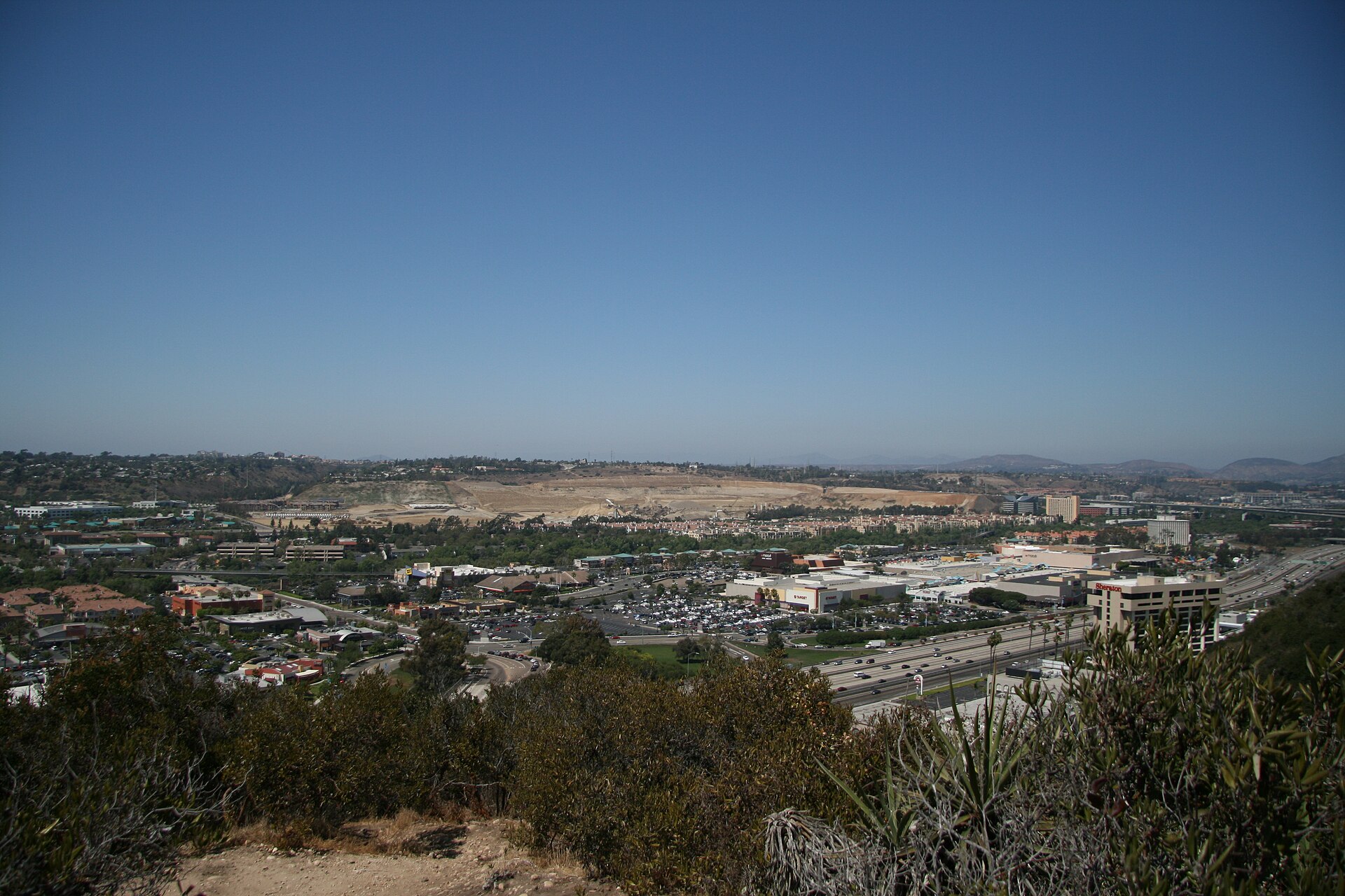 A wide view across central Mission Valley in San Diego from a brushy hillside, with buildings and freeways on the valley floor under a clear blue sky