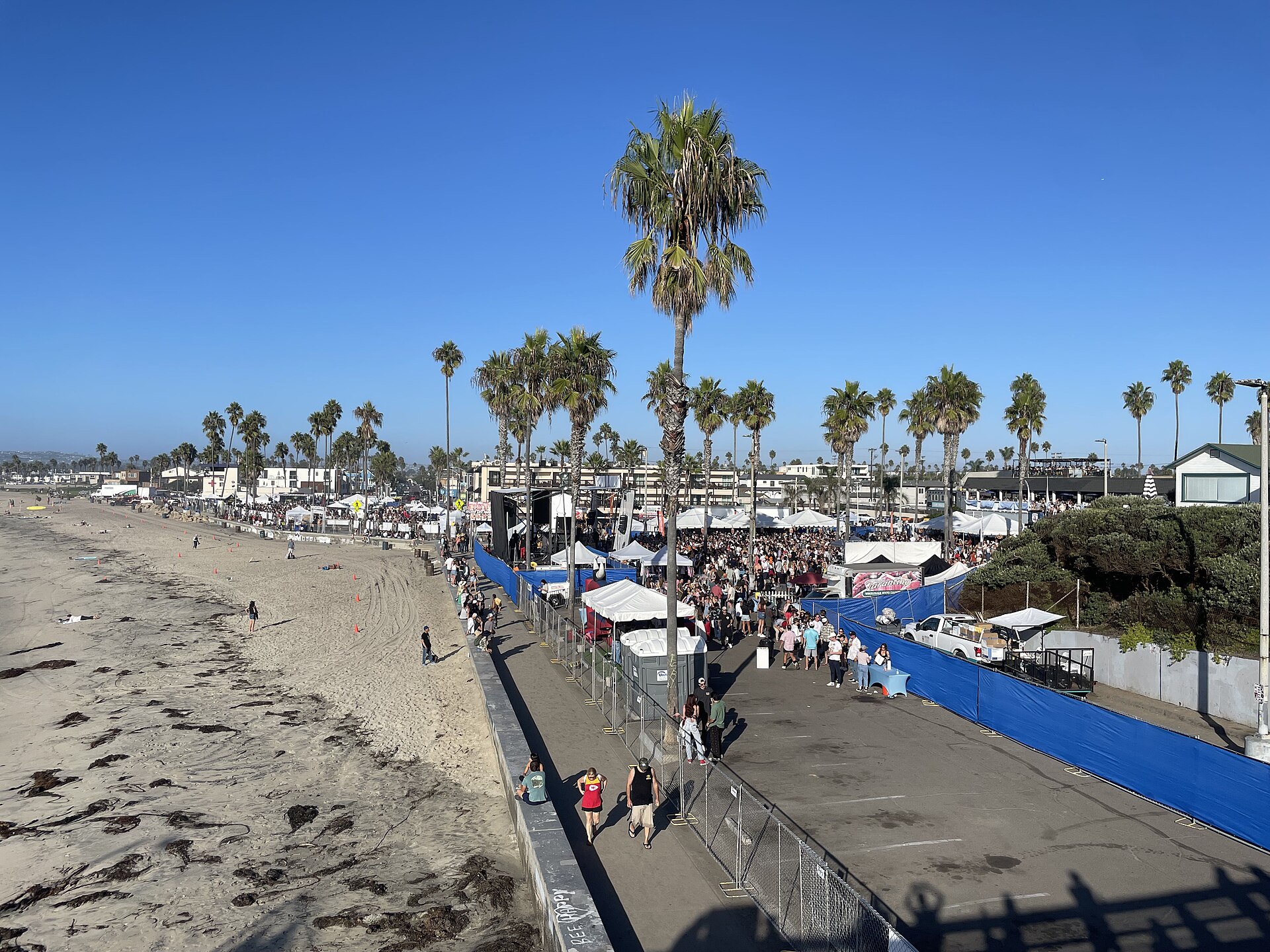 Festival tents and crowds along the beachfront parking lot at OB Oktoberfest 2025, seen from the Ocean Beach Pier, with palm trees and blue sky