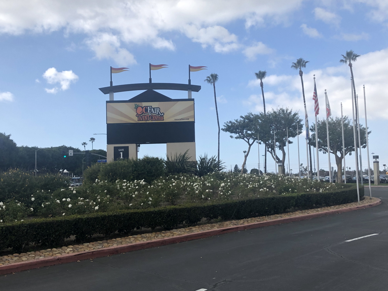 The entrance sign of the OC Fair and Event Center in Costa Mesa, with palm trees, flags, and landscaped shrubs along the road