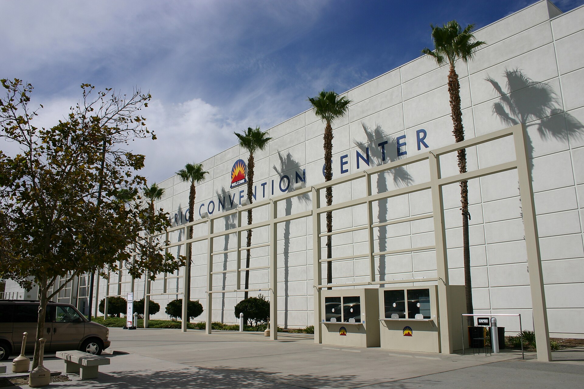 The white facade of the Ontario Convention Center with its name in large letters, lined with tall palm trees under a partly cloudy sky