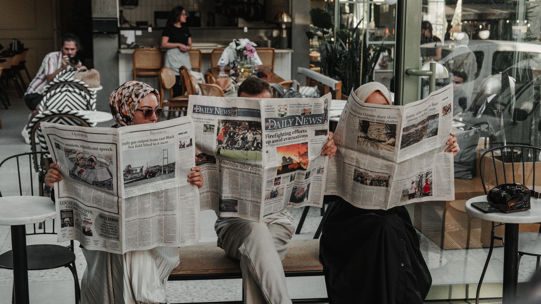 Three women reading newspapers at a cafe table.