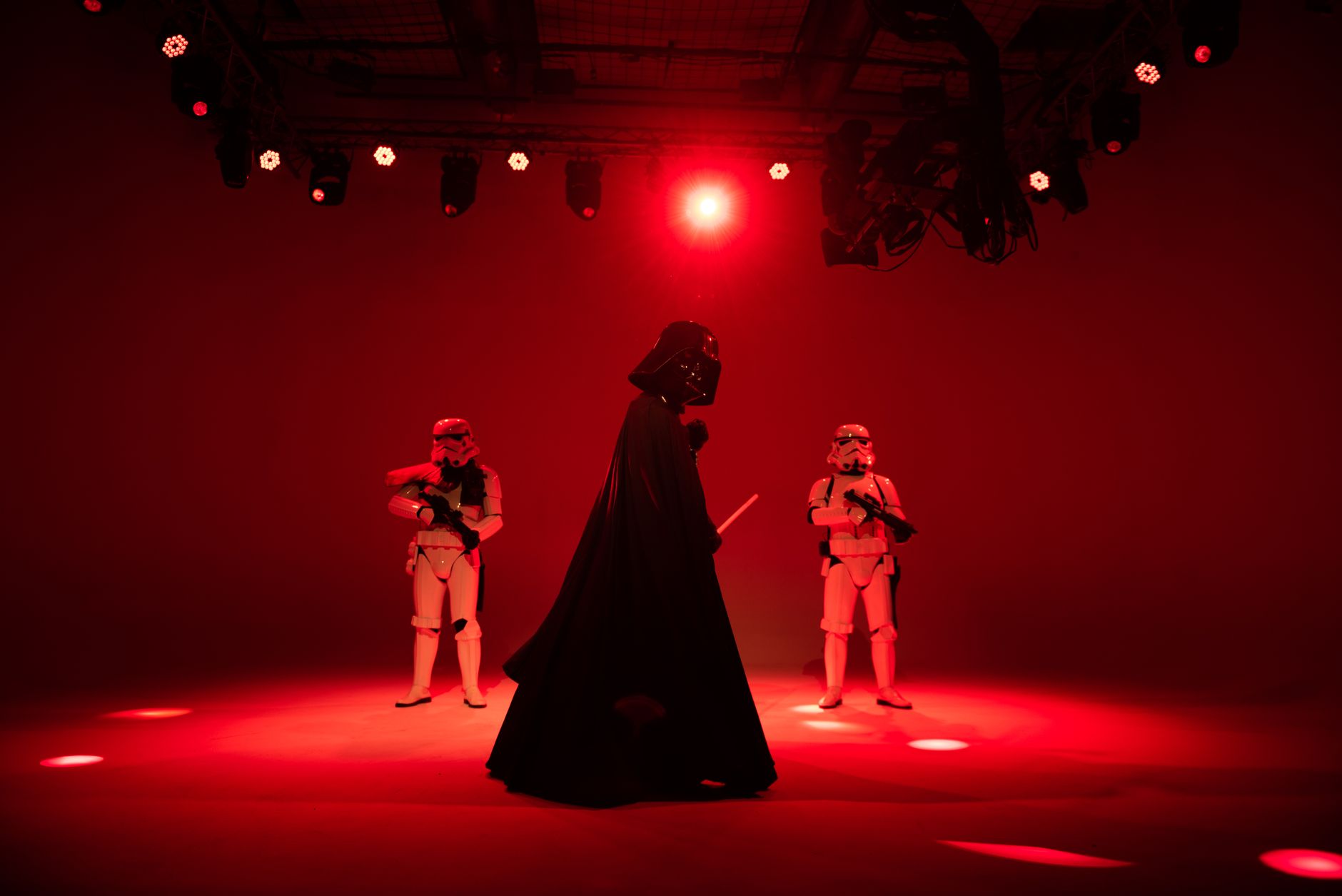 Darth Vader stands center stage in a flowing cape, flanked by two stormtroopers under intense red stage lighting.