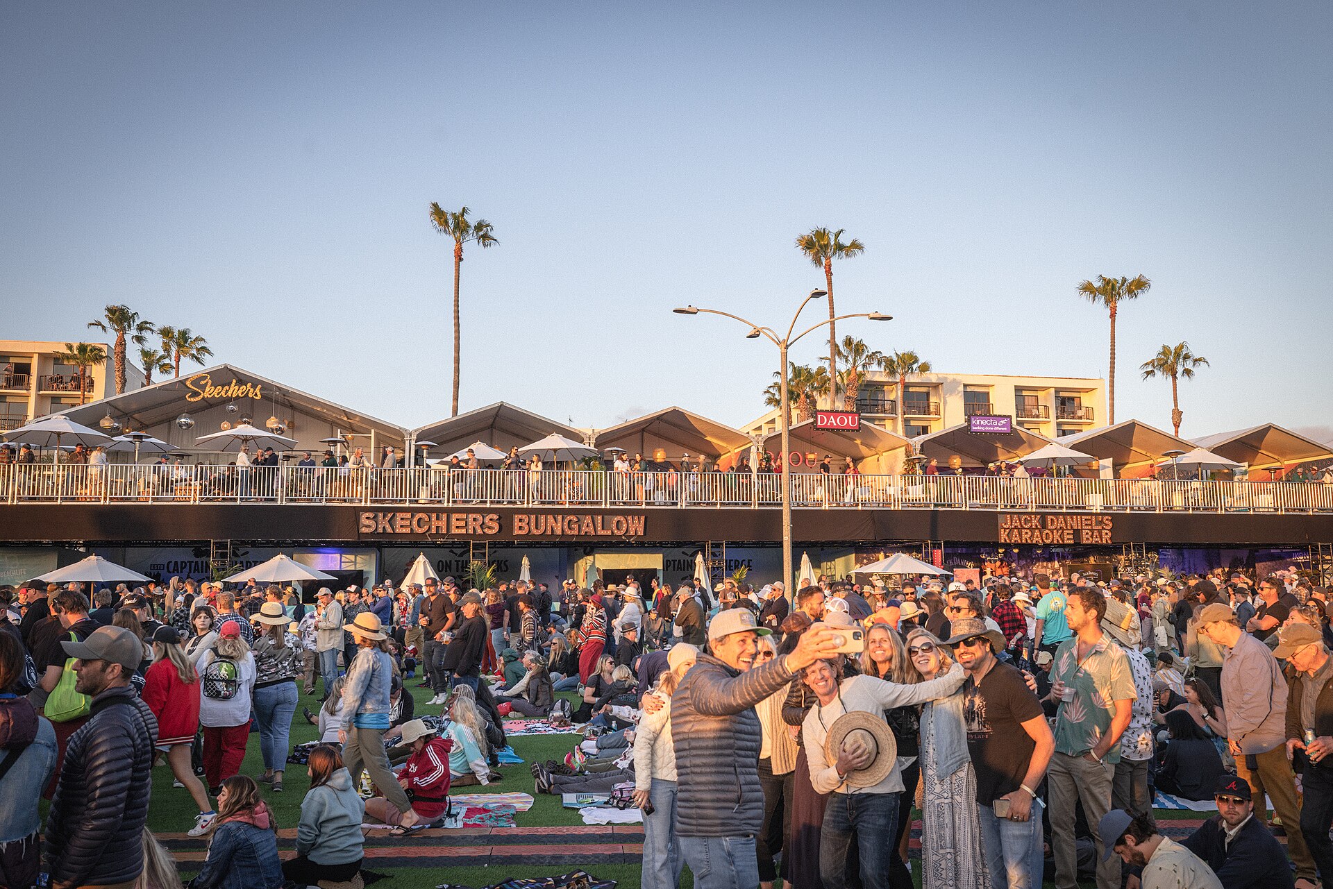 A daytime festival crowd in front of two-level festival structures and palm trees on the Redondo Beach waterfront