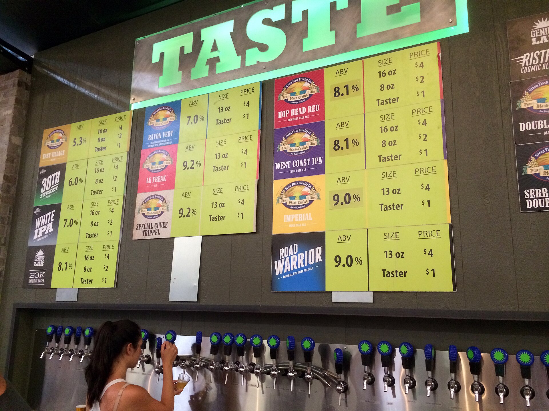 A bartender pulls a beer from a long row of taps under a bright green TASTE sign and chalkboard beer menus at a San Diego brewery