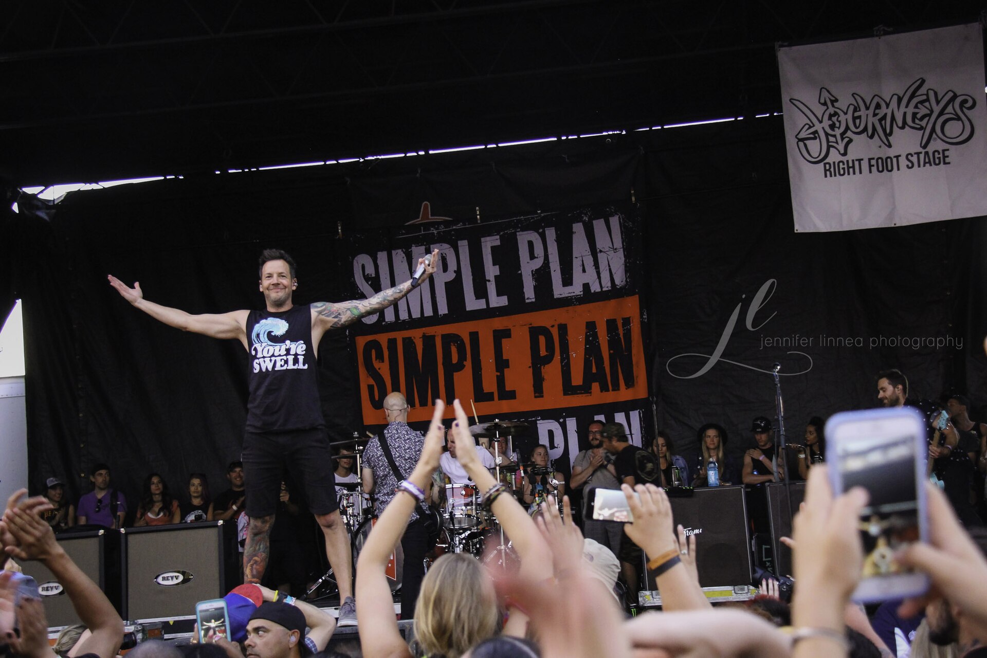 Simple Plan's singer onstage with arms spread wide in front of a Simple Plan banner as fans raise their hands
